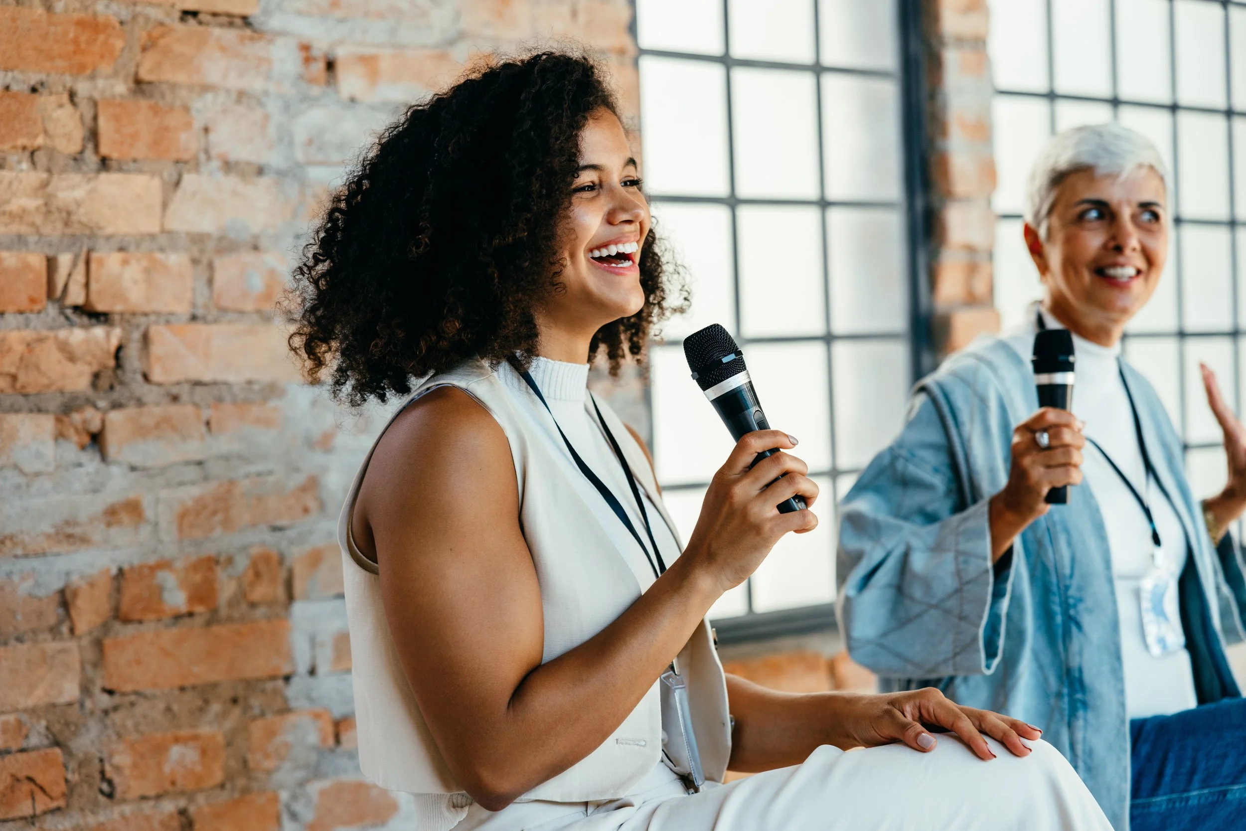 Two women speaking into handheld microphones during a panel discussion or presentation in front of brick wall and large window.