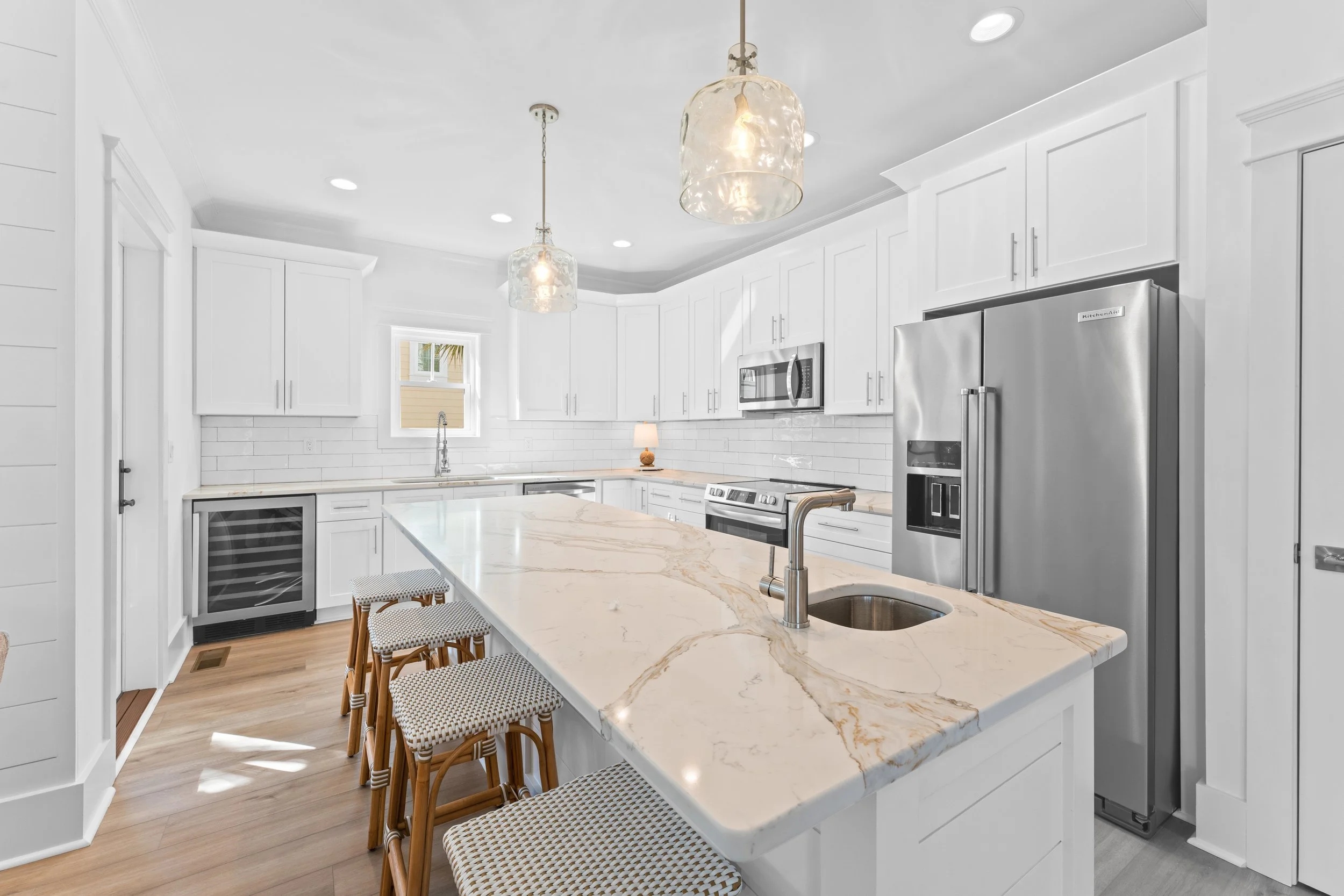 Modern white kitchen with marble island, stainless steel appliances, hanging pendant lights, and light wood flooring.