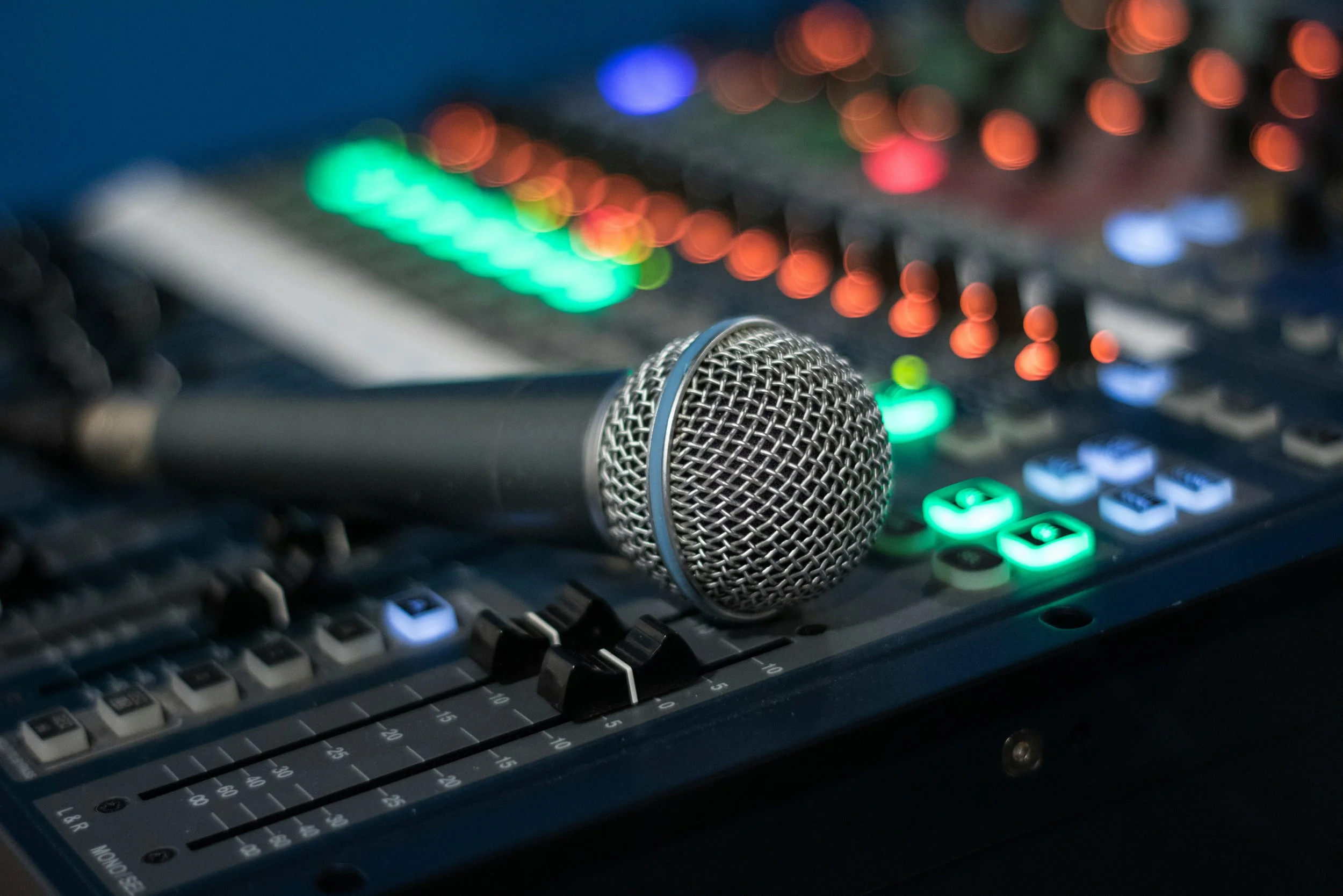 A microphone resting on a sound mixing console with colorful illuminated buttons and sliders, used for live sound or recording.