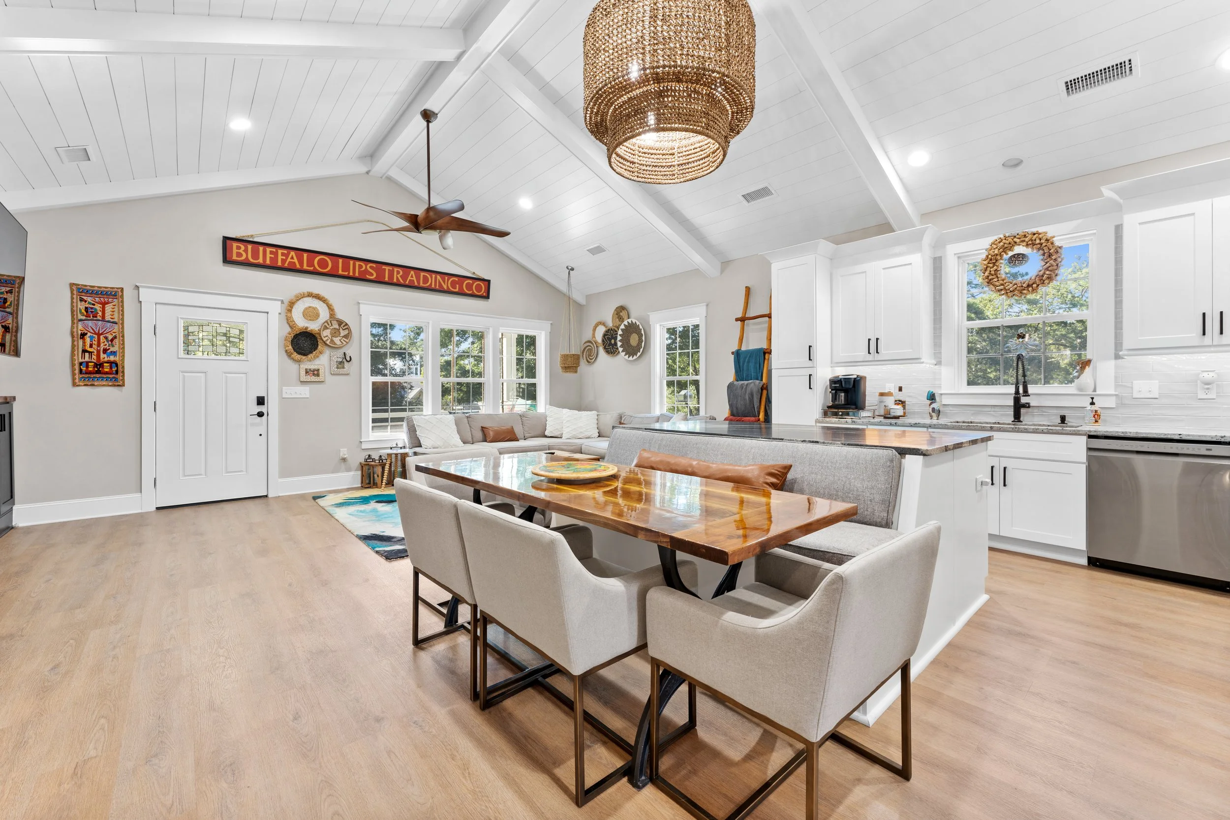 Open-concept kitchen and living room with white cabinetry, a wooden dining table with chairs, a beige sofa, artwork, and decorative items, with large windows and a white vaulted ceiling.