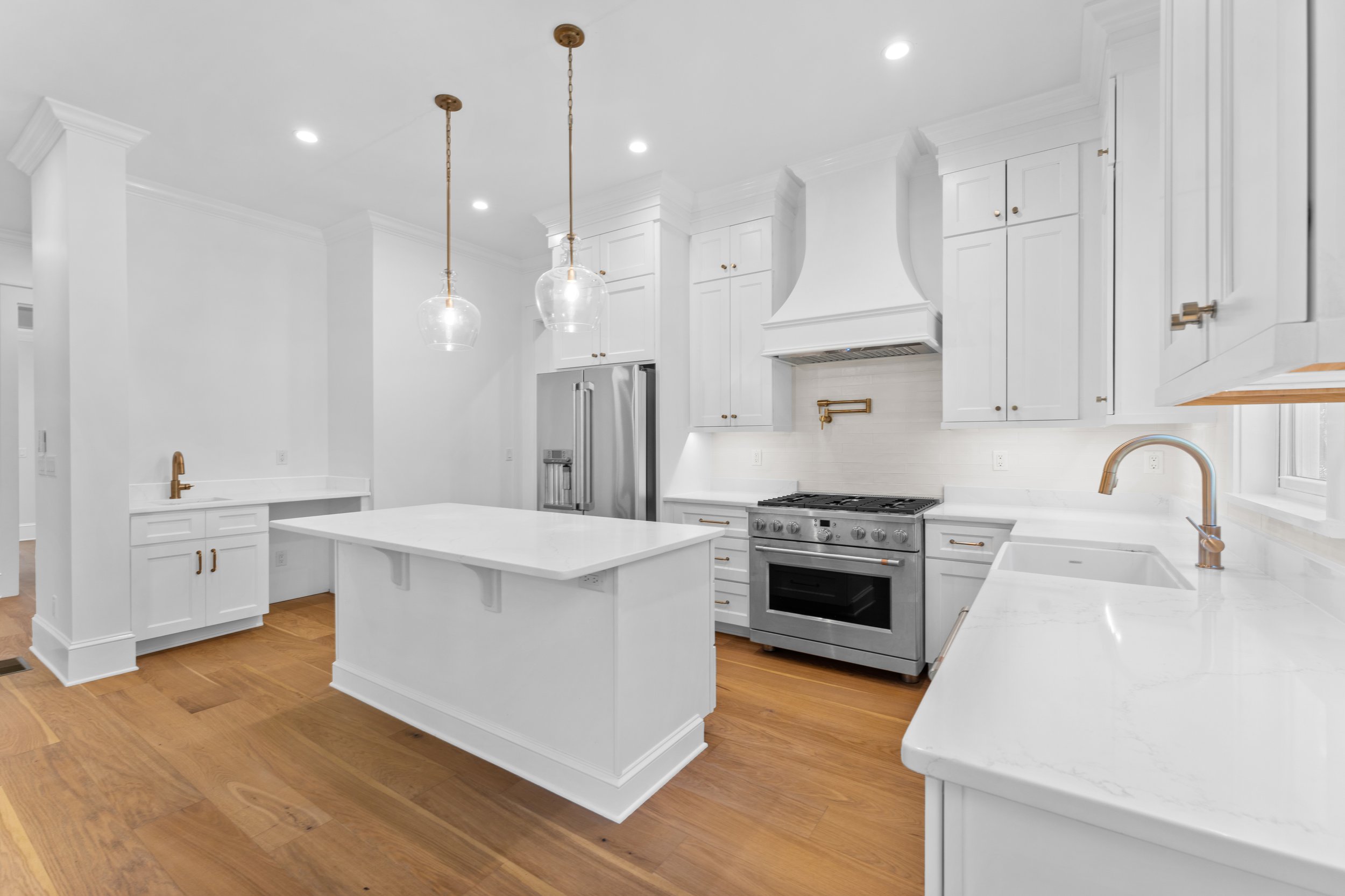 Modern white kitchen with wooden floors, white cabinets, a kitchen island, stainless steel appliances including a refrigerator and oven, and brass fixtures including pendant lights and faucets.