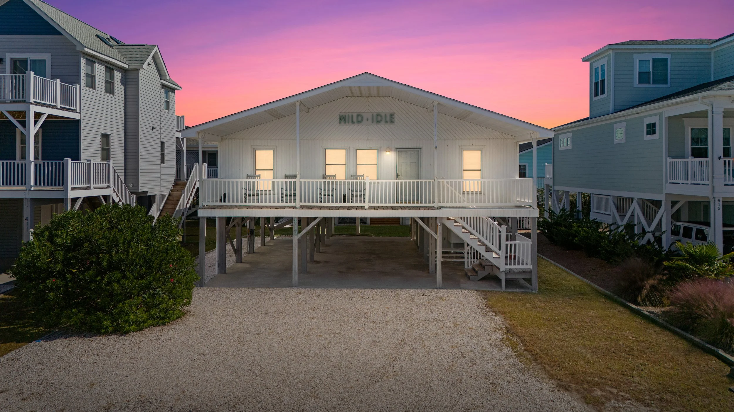 Beach house with white exterior on stilts, under a colorful sunset sky, surrounded by other similar homes, with a gravel driveway and landscaped bushes in the front.