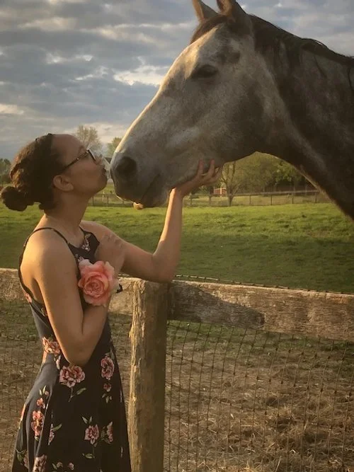A woman in a floral dress, holding a pink rose, kisses a large gray horse on its nose in a fenced outdoor field during sunset.