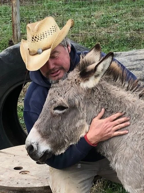 A man wearing a cowboy hat hugs a gray donkey.