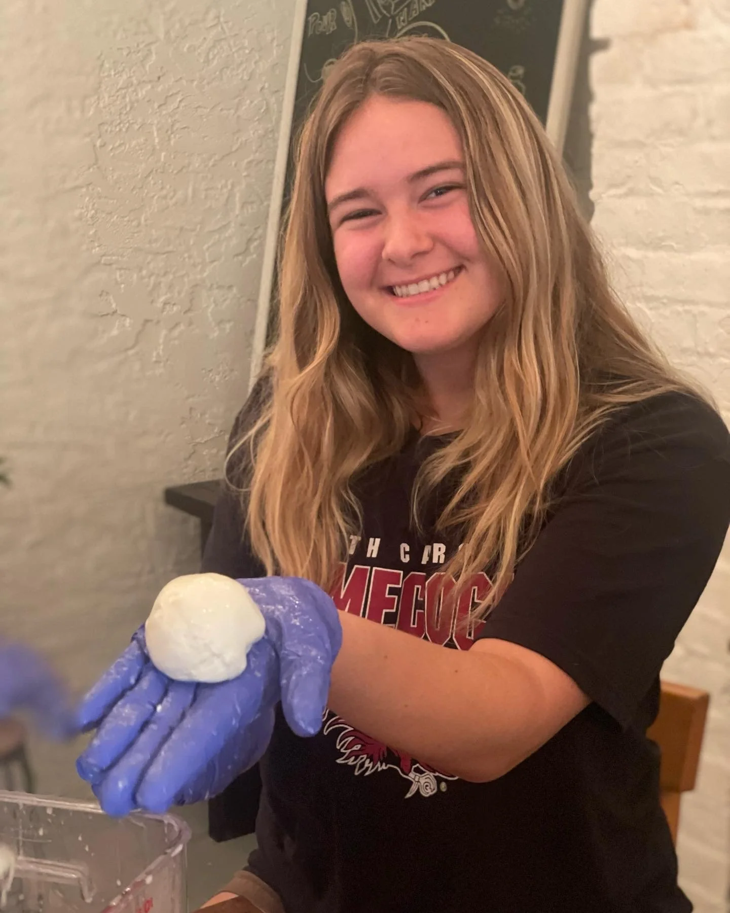 A smiling young woman with long wavy blonde hair wearing a black T-shirt with red text holds a ball of dough covered with flour in her gloved hand.