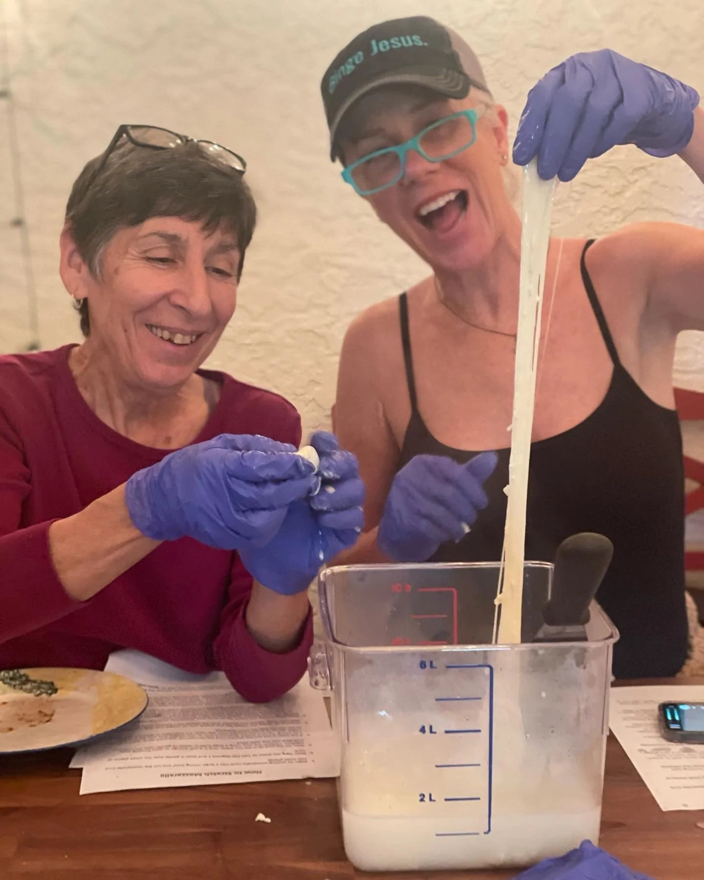 Two women with blue gloves preparing cheese in a restaurant. They are smiling and enjoying the activity, with one pulling cheese and the other holding a small tool.