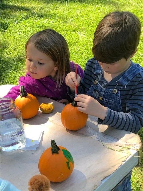 A young girl and boy are sitting at a table outdoors, painting small pumpkins. The girl is wearing a pink jacket and the boy is wearing a striped shirt with denim overalls. There is a jar of water, a piece of paper towel, and a few painted pumpkins o