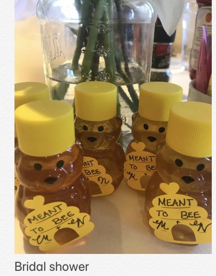 Several small bear-shaped containers with yellow caps and tags that read 'Meant to Bee' are arranged in front of a clear vase of green stems. The containers likely hold honey for a bridal shower.