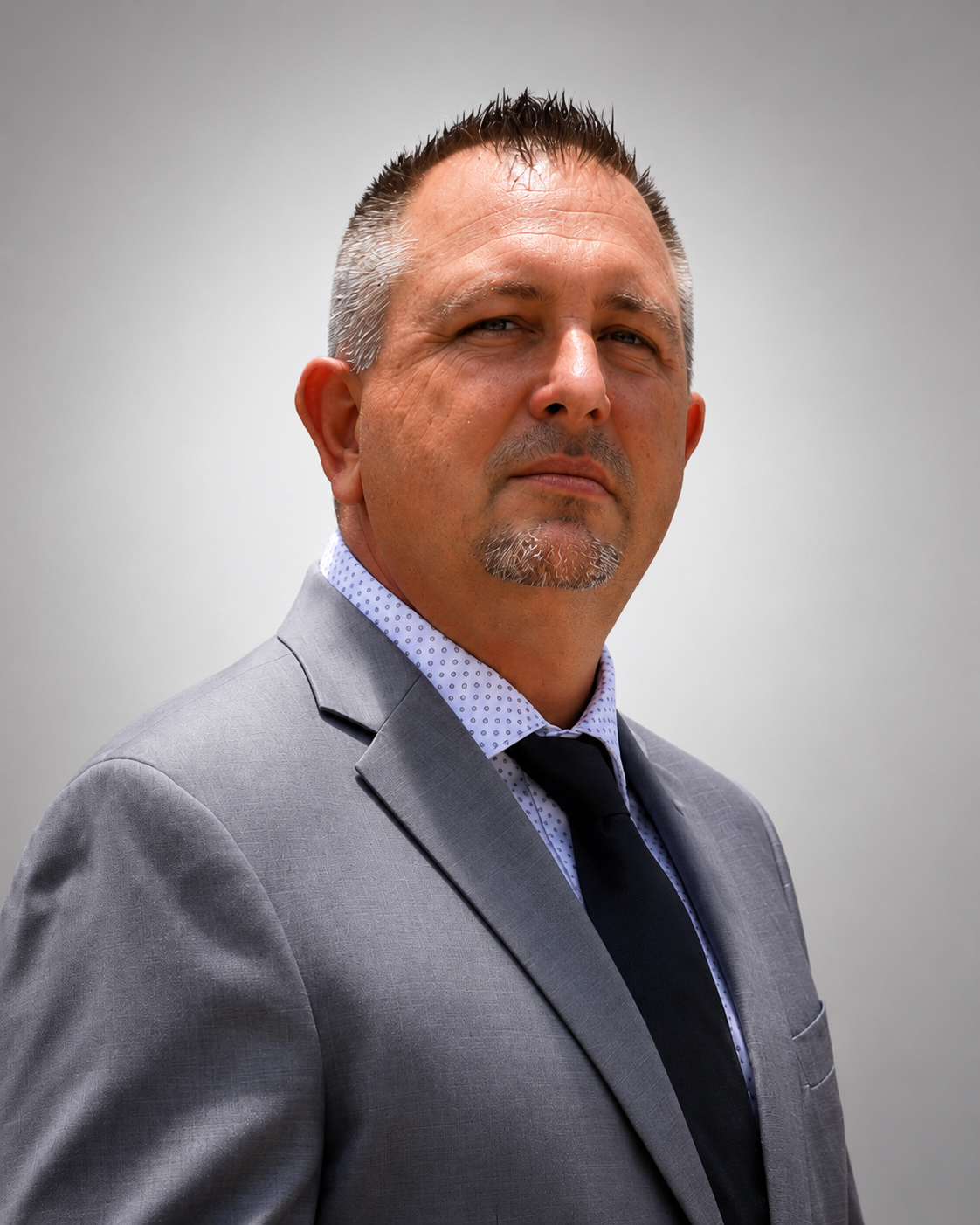 A middle-aged man in a gray suit and black tie, standing against a plain light gray background.