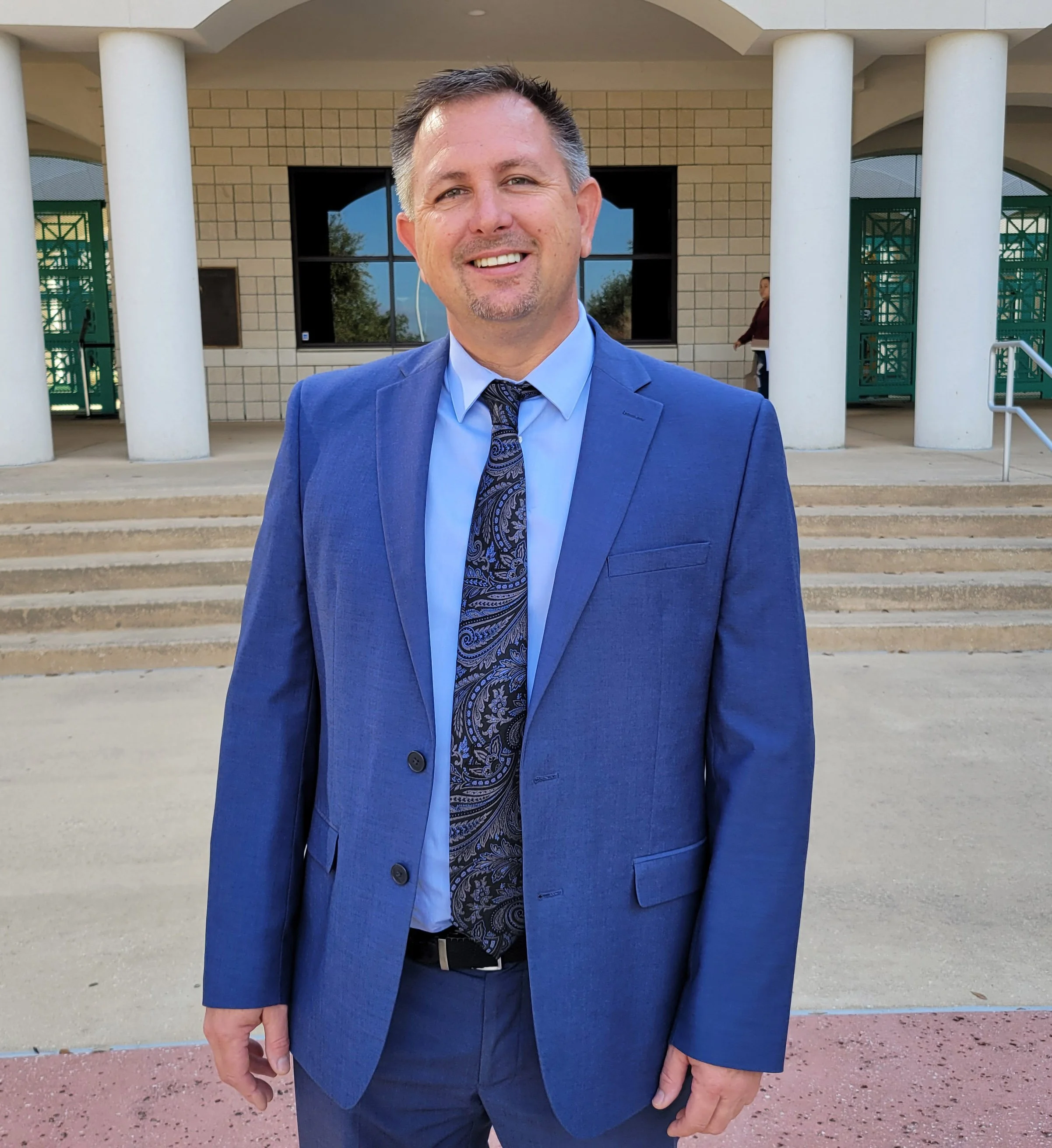 A man in a blue suit and patterned tie stands outside a building with white columns and brick walls, smiling at the camera.