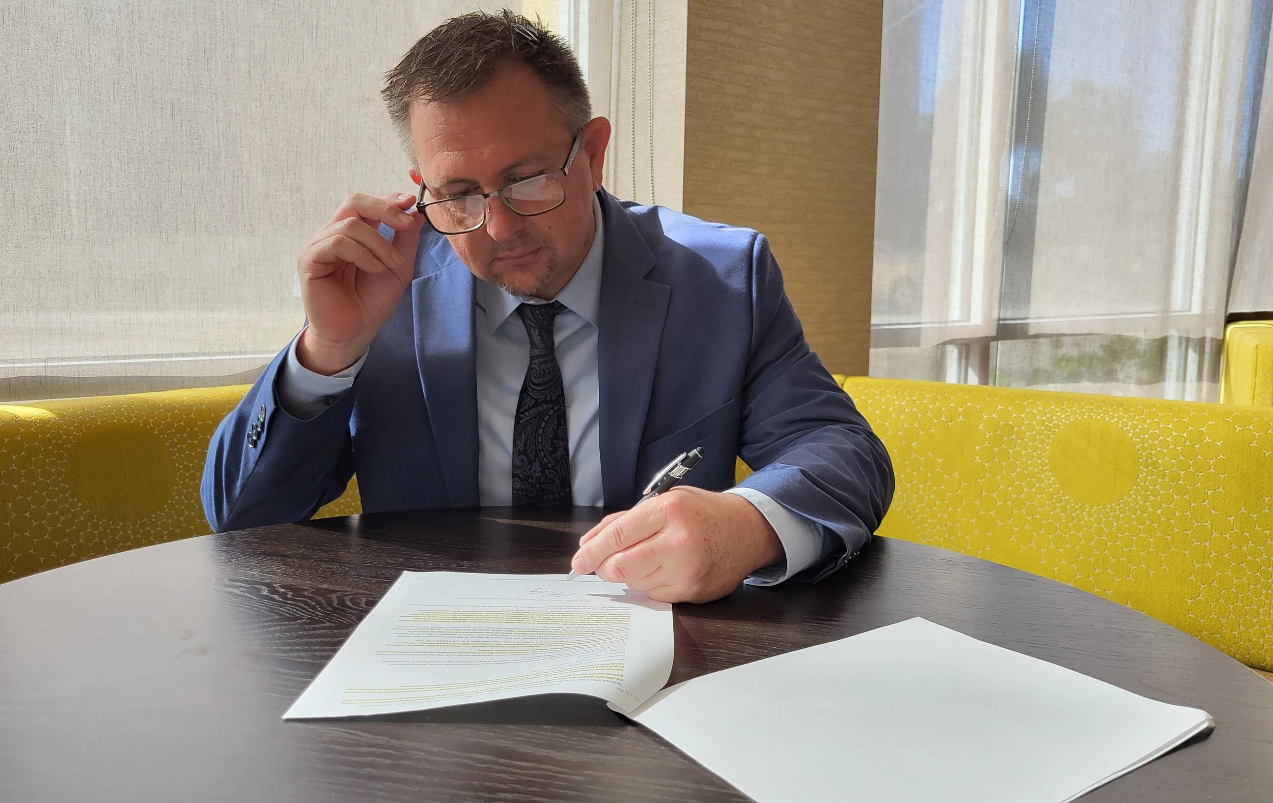 A man in a blue suit and patterned tie is sitting at a table, reading and marking pages in a document. He is wearing glasses and is adjusting them with his left hand, while holding a pen in his right hand. Yellow chairs are behind him near large windows with sheer curtains.