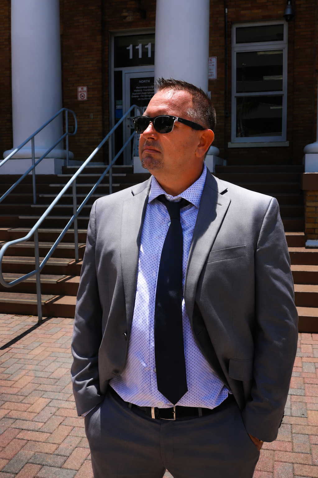 A man in a gray suit, white shirt with small blue polka dots, black tie, and sunglasses standing outdoors in front of a building with steps, columns, and the number 111 above the door.