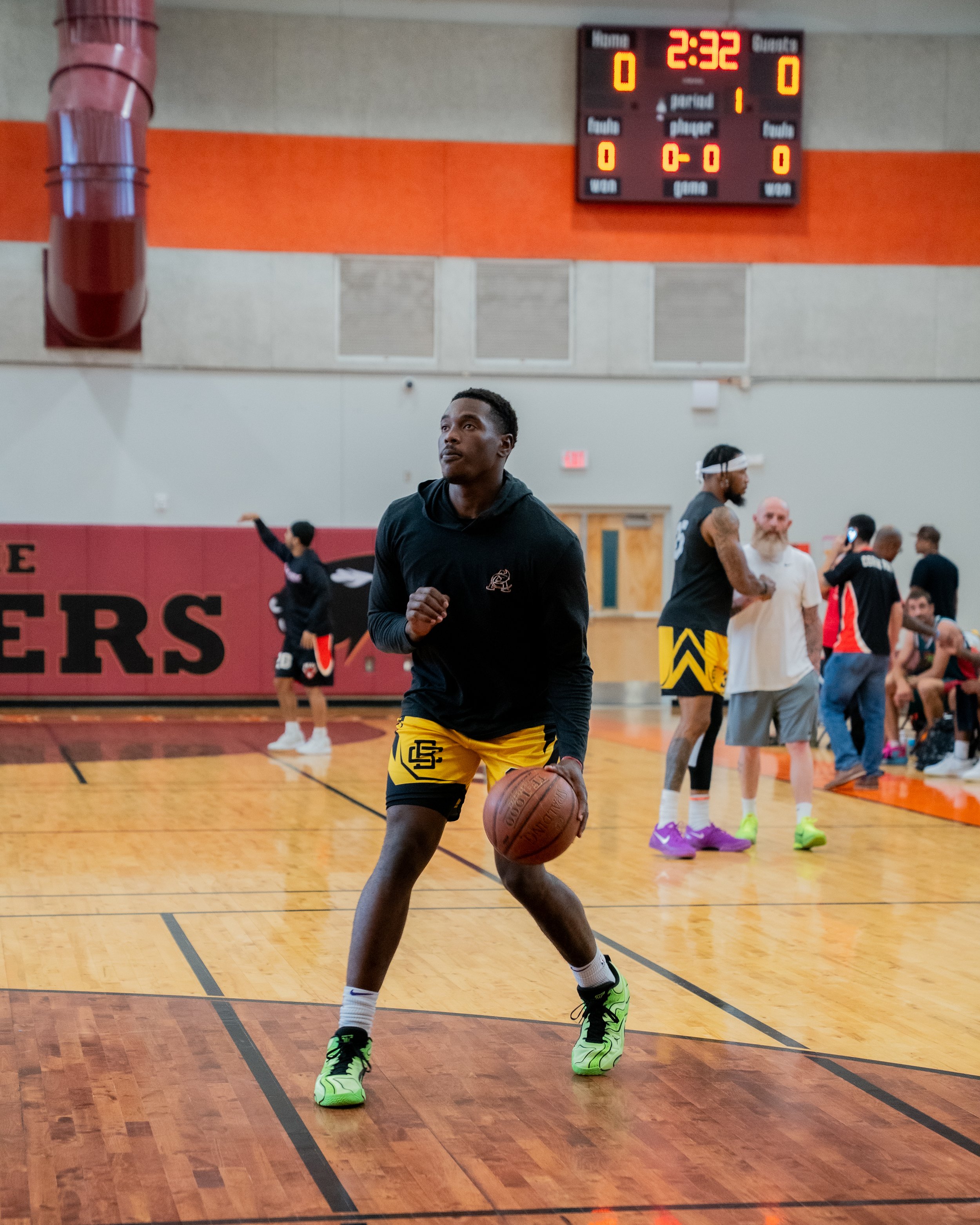 A young man practicing basketball on an indoor court, dribbling a basketball, while wearing a black hoodie, yellow shorts, and bright green sneakers. In the background, other players and spectators are visible, with a digital scoreboard showing 2:32 remaining in the game.