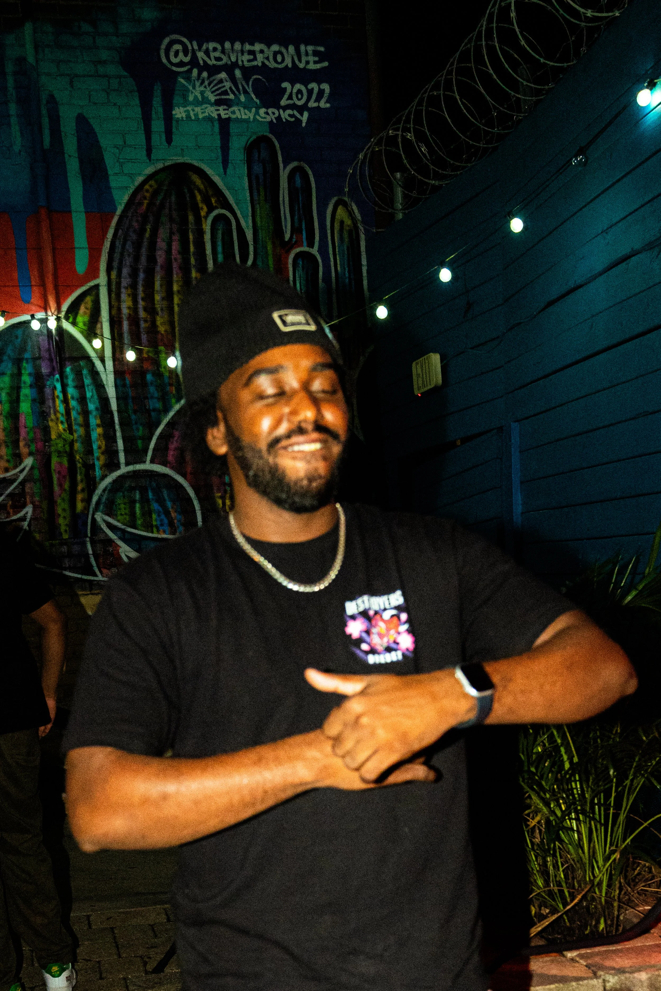 A man with a beard and mustache wearing a black beanie, black T-shirt, chain necklace, and smartwatch, smiling with his eyes closed in front of a colorful graffiti mural and string lights at night.