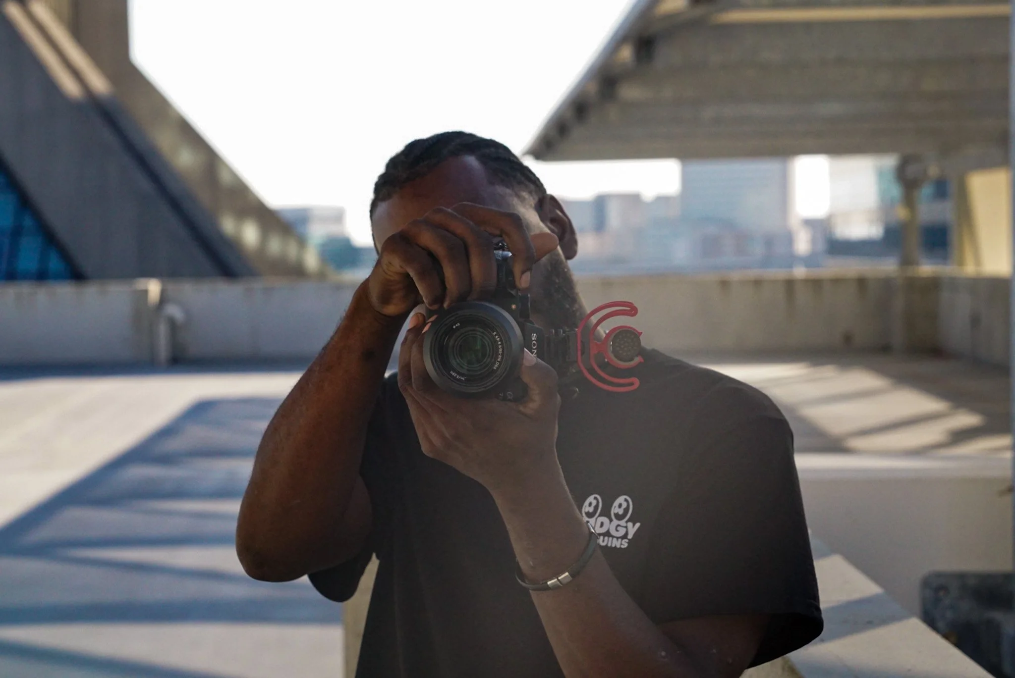 A person takes a photo of a reflection with a camera on a rooftop parking lot, with a cityscape in the background.