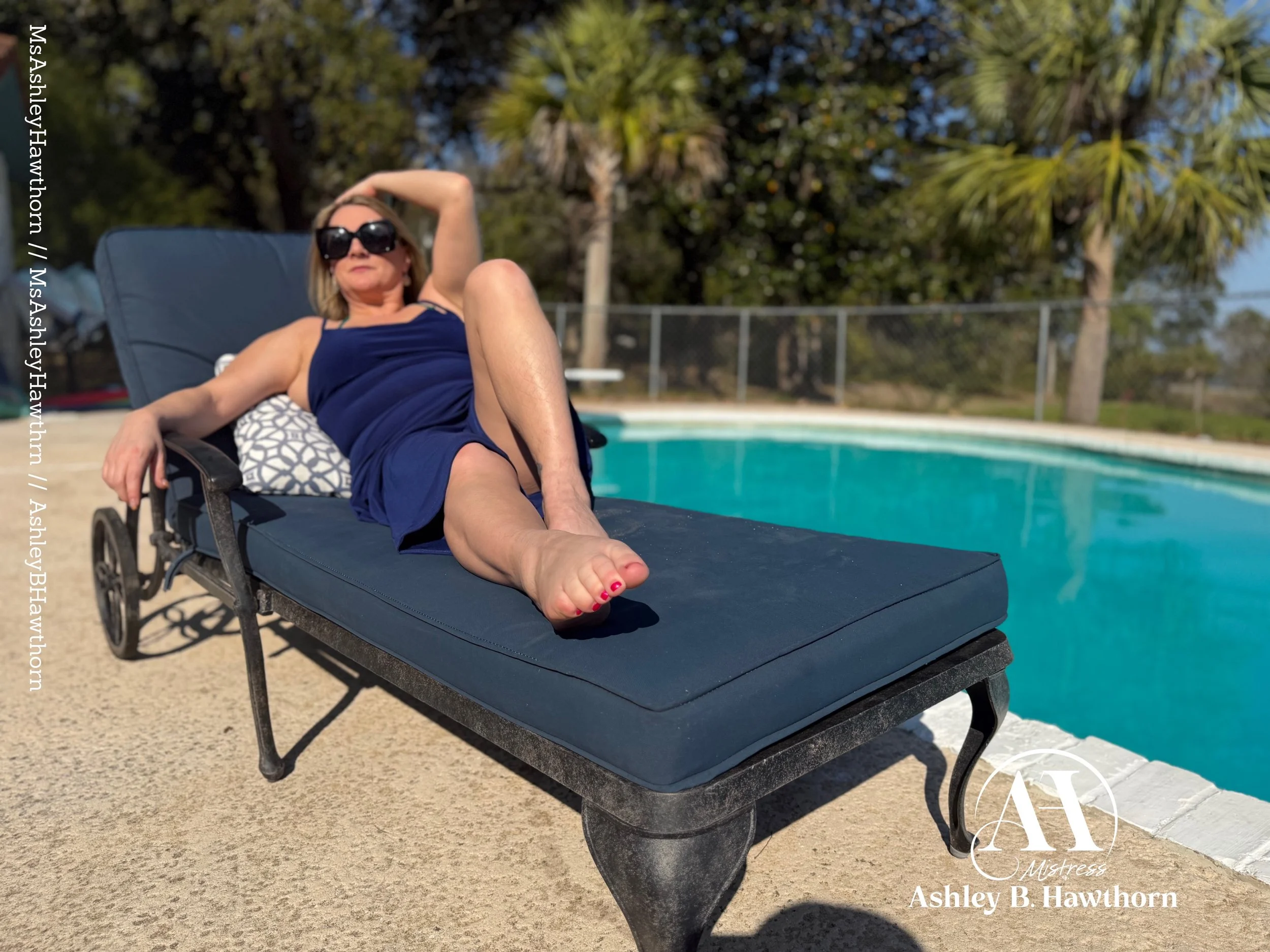 Woman relaxing on a lounge chair by a swimming pool surrounded by trees, wearing sunglasses and a blue dress.