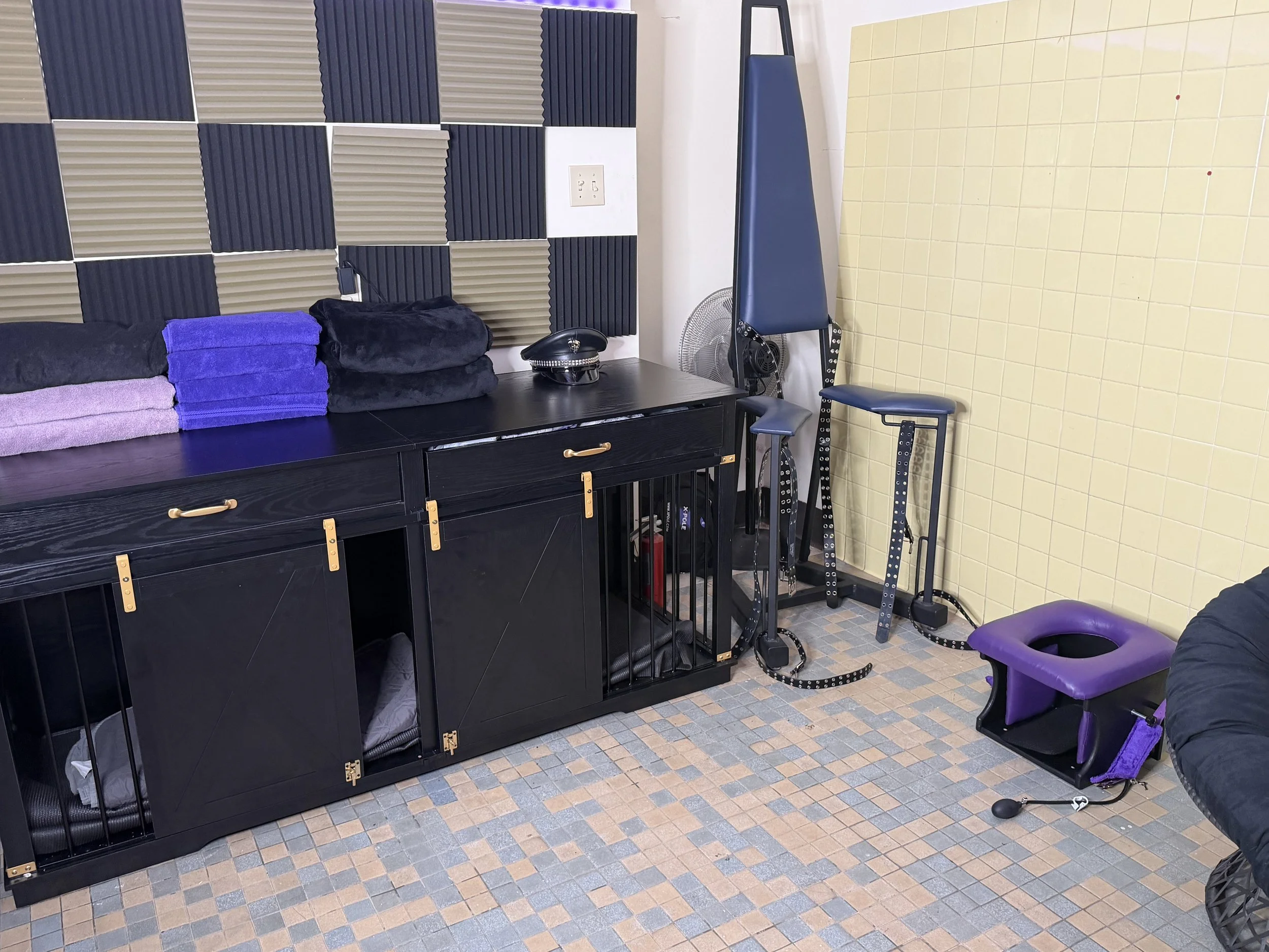 A room with soundproofing foam panels on the wall, containing black, gray, and cream-colored sections. There is a black cabinet with a golden handle and latch, several folded towels, and a black helmet on top. To the right, there is a blue exercise bench, a fan, and a purple chair. The floor has small checkered tiles in shades of gray and beige.