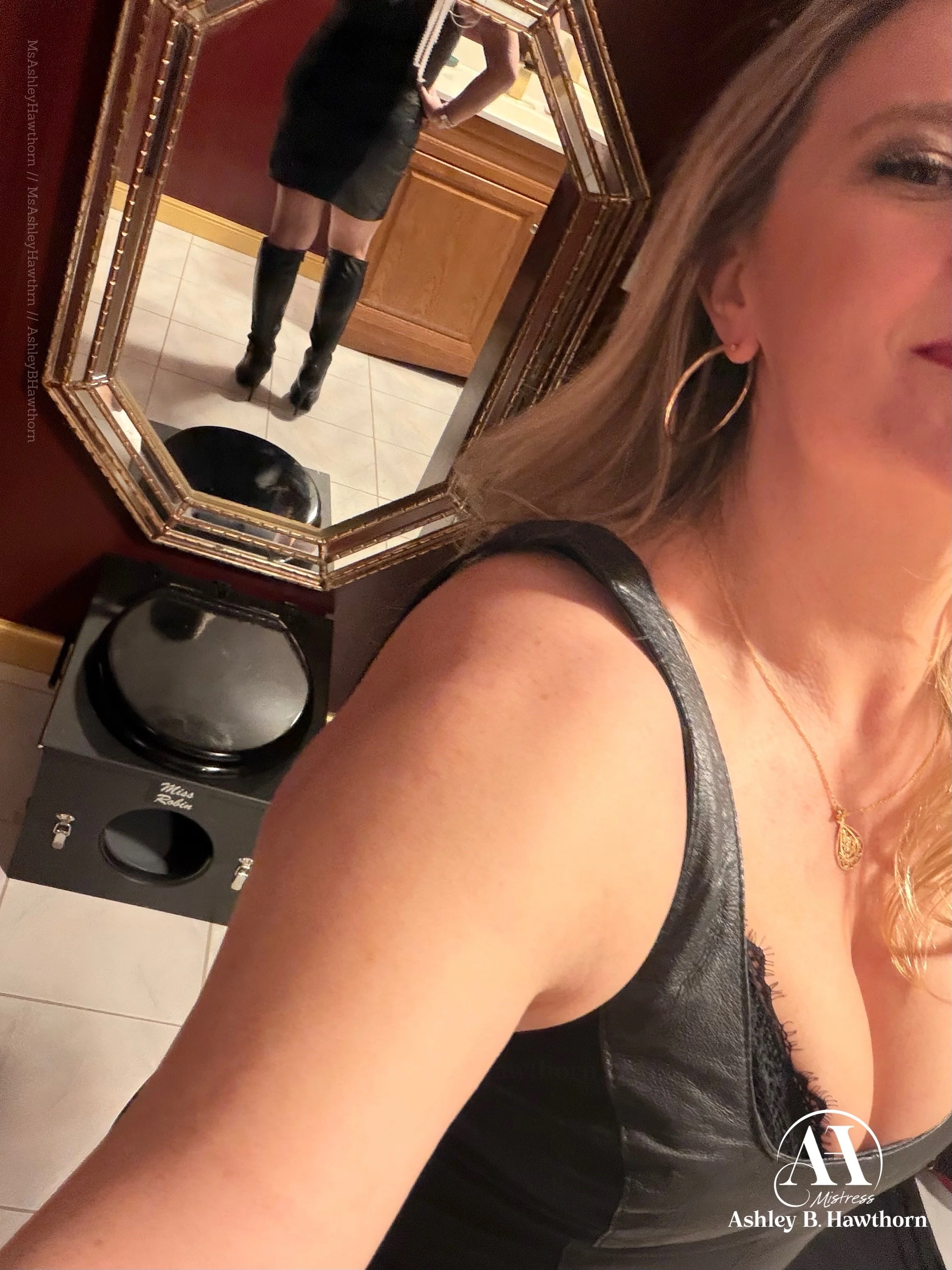 A woman wearing black leather boots, a black dress, and large hoop earrings is taking a mirror selfie with her phone, capturing her lower body and part of her face with long, wavy hair. The scene is set indoors with red walls, wooden cabinets, and a 