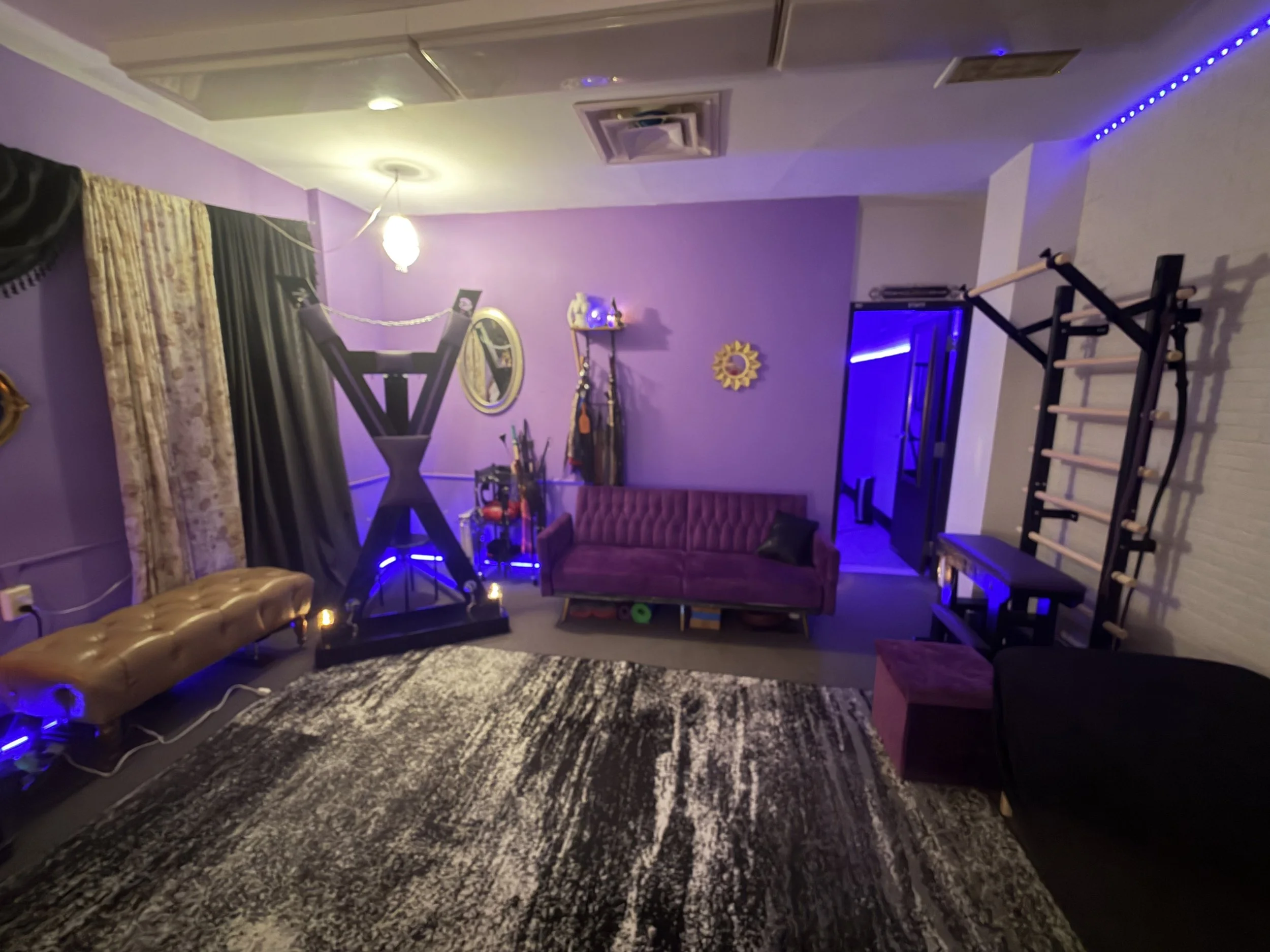 View of a cozy room with purple walls, a vintage-style sofa, a patterned rug, exercise equipment, and decorative mirrors illuminated by soft purple and yellow lighting.