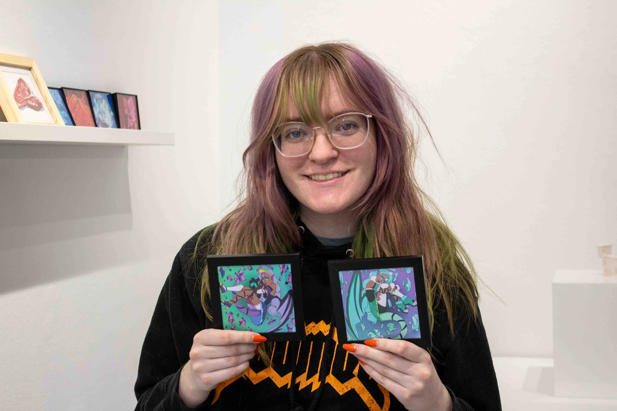 Student artist Callum Landrum stands in the Shop Columbia display titled “MINI MINI: A Showcase of Miniature Works” holding their pieces titled, “Gem Dragon Green” and “Gem Dragon Purple” during the artist reception on Thursday, Apr. 9, 2026. 