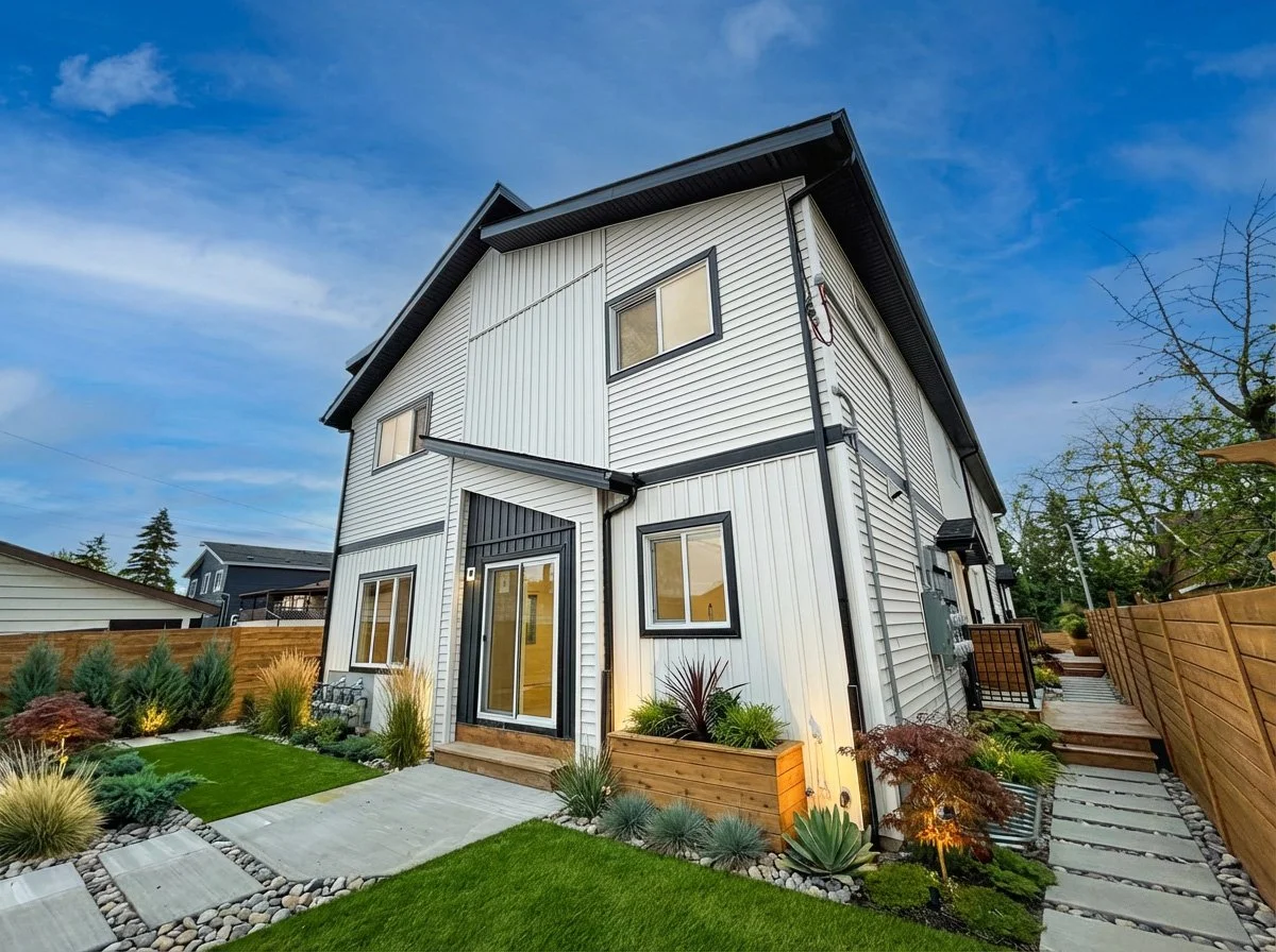 Modern white two-story house with black trim, landscaped front yard with grass, stone pathway, and plants, under a blue sky during daytime.