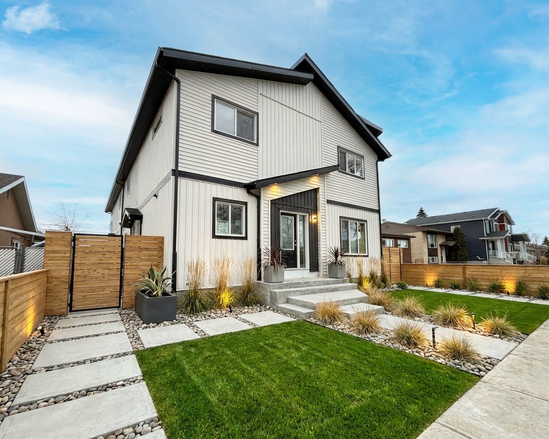 Modern two-story house with white siding and black trim, small front porch with steps, landscaped yard with grass, shrubs, and pathway, wooden fences on sides, and neighboring houses in background under a partly cloudy sky.
