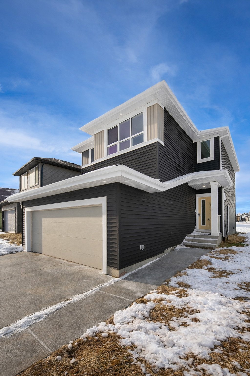 A modern two-story house with black siding, white trim, a beige front door, a attached garage, steps leading to the entrance, and partially snow-covered front yard under a blue sky.