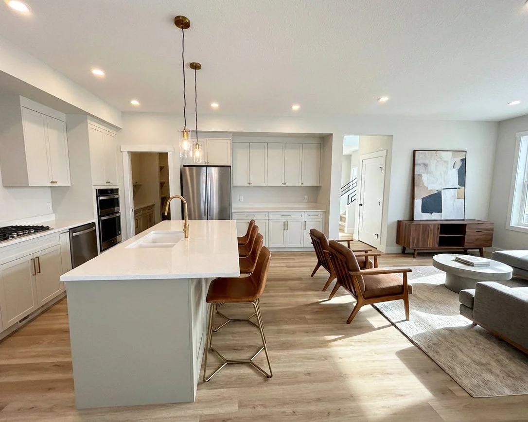 Open concept kitchen and living room with white cabinets, stainless steel appliances, a large kitchen island, wooden chairs, a gray sofa, and a modern wooden media console, with natural light coming from windows.