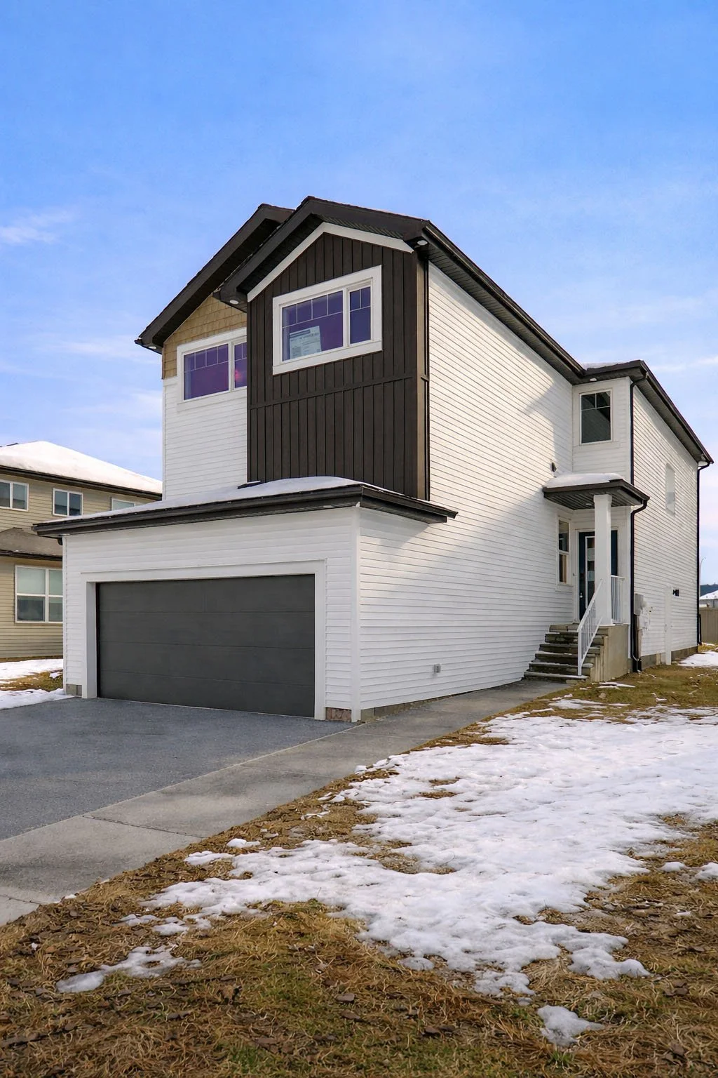 A modern two-story house with white and dark brown exterior siding, a gray garage door, and steps leading to the front door. The yard has patches of snow on the ground under a clear blue sky.