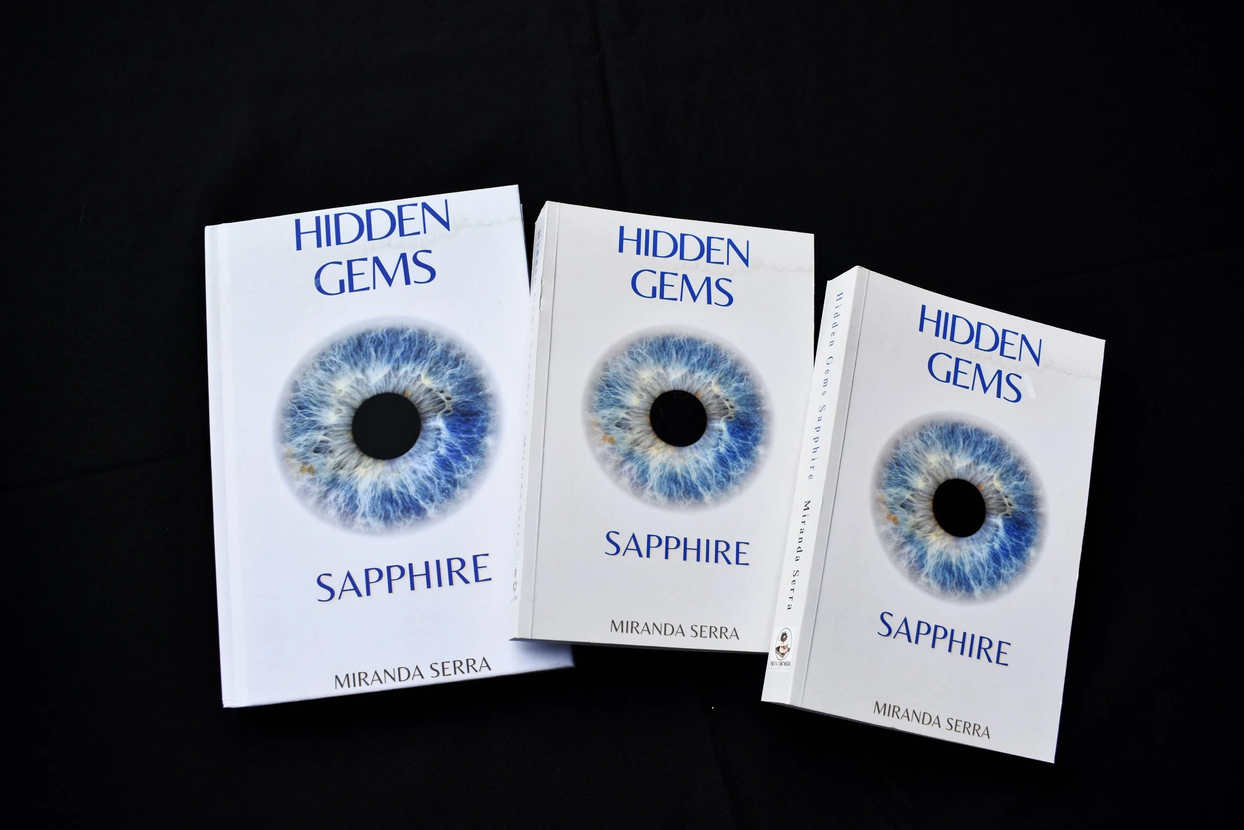 Three copies of the book titled "Hidden Gems Sapphire" by Miranda Serra, displayed against a black background. The books feature a cover with a detailed image of a blue and white eye, with the title and author's name printed on the cover.
