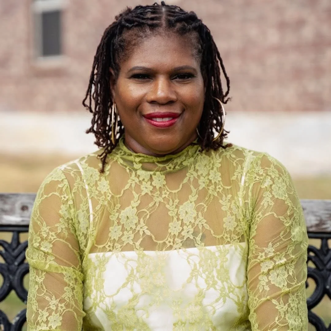 A woman with dark braided hair and earrings, wearing a yellow lace top, is sitting on a bench outdoors with a blurred brick building in the background.