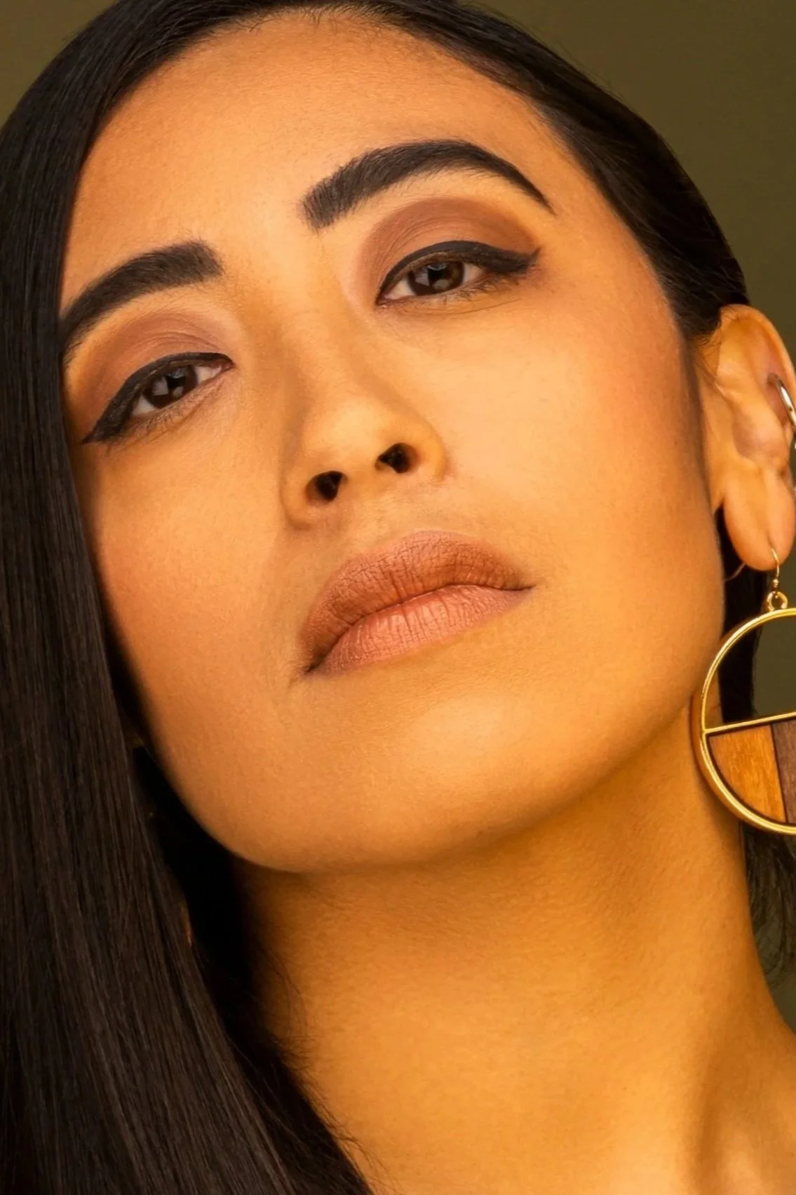 Close-up portrait of a woman with long black hair, wearing makeup with bold eyeliner and matte lipstick, and gold geometric earrings.