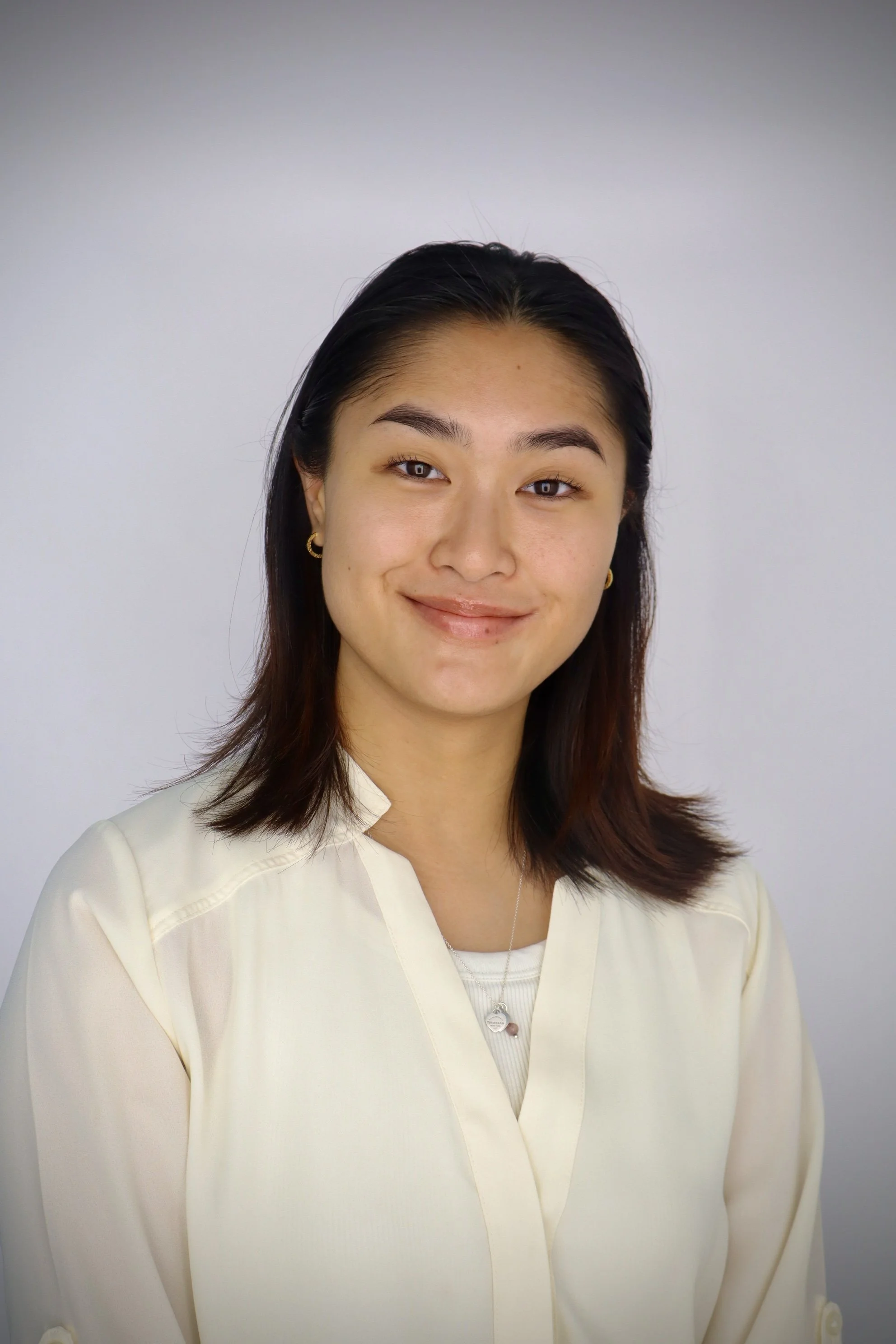 A young woman with dark brown hair, wearing a cream-colored top, smiling at the camera against a plain light gray background.