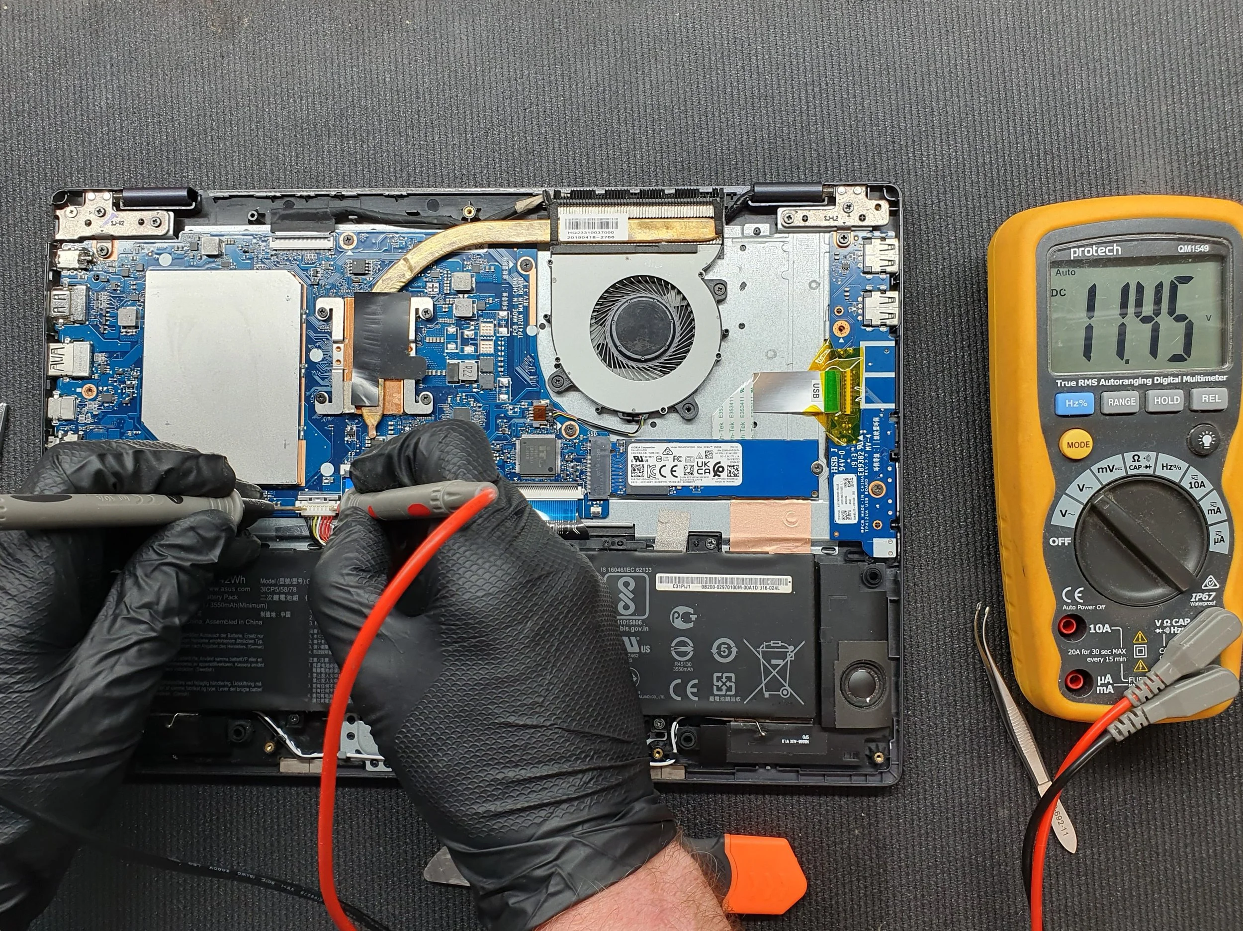 A person wearing black gloves is using a multimeter with red and black probes to test a laptop's circuitry. The laptop has its bottom cover removed, exposing the internal components, including the battery, motherboard, and cooling fan. The multimeter readings show 11.45 volts.
