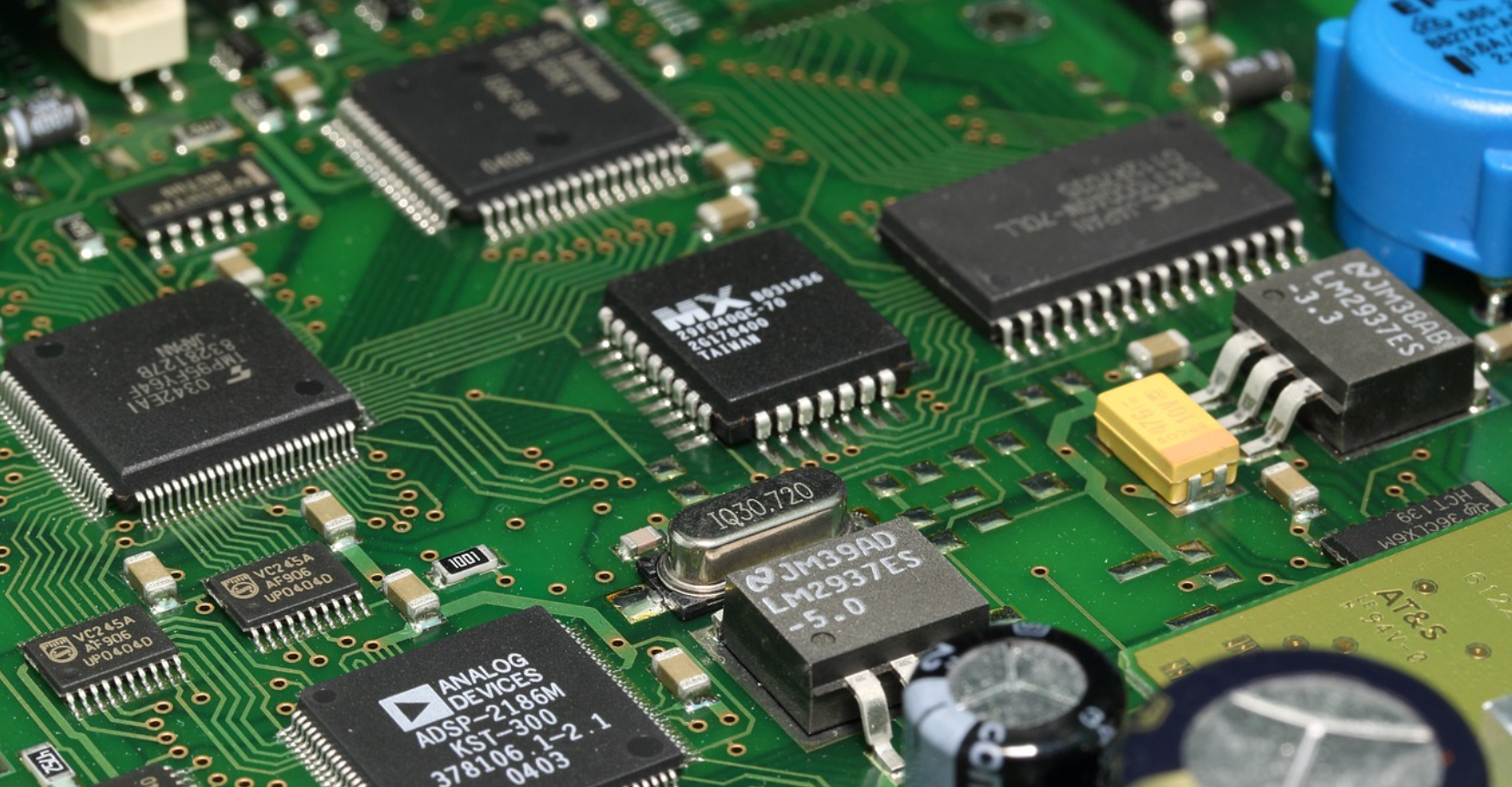 Close-up of a green printed circuit board (PCB) with various electronic components including integrated circuits, capacitors, resistors, and connectors.