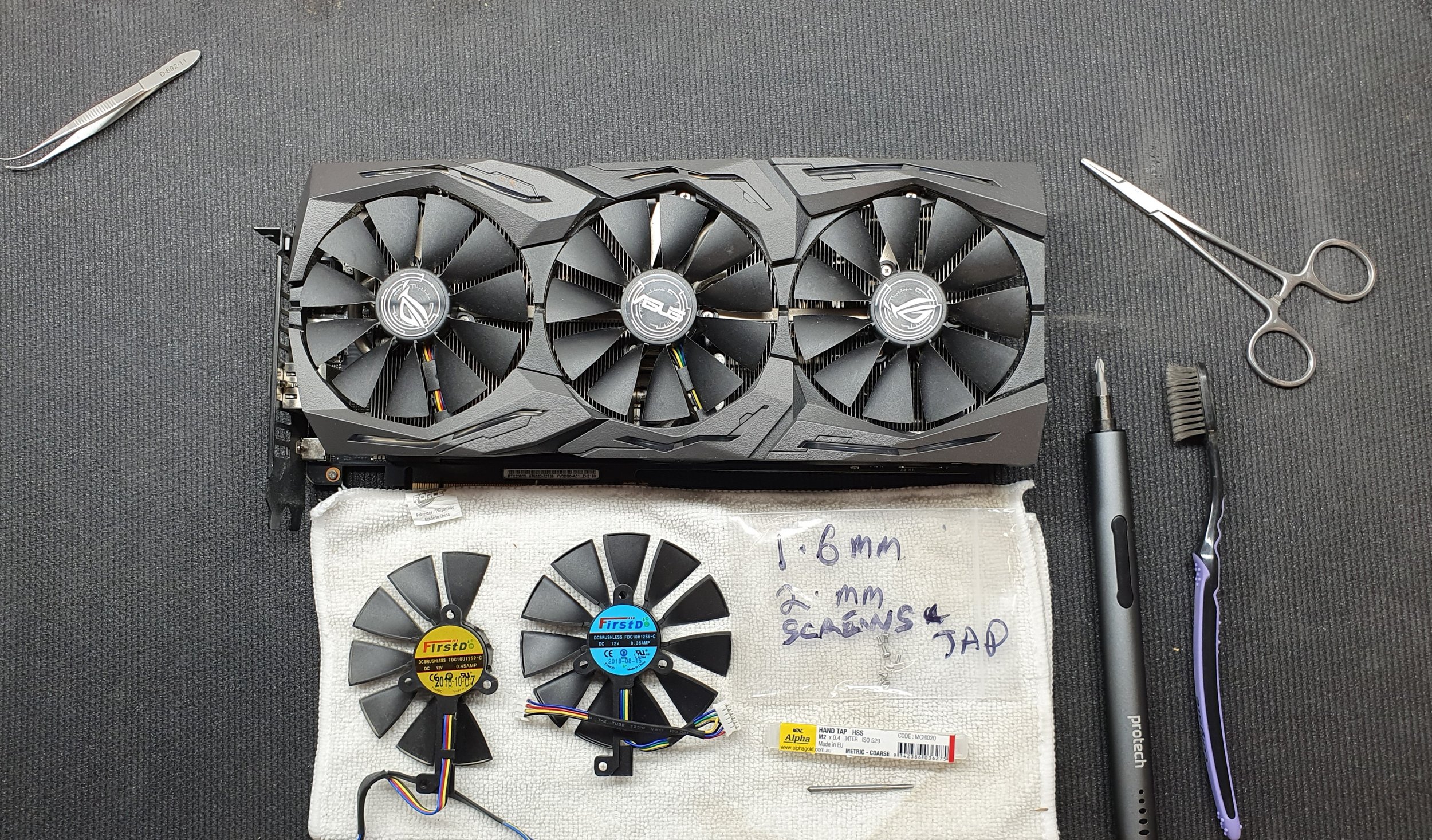 A large graphics card with three cooling fans, two smaller cooling fans, a screwdriver, tweezers, and scissors on a black textured surface, with handwritten notes on paper beneath the smaller fans.