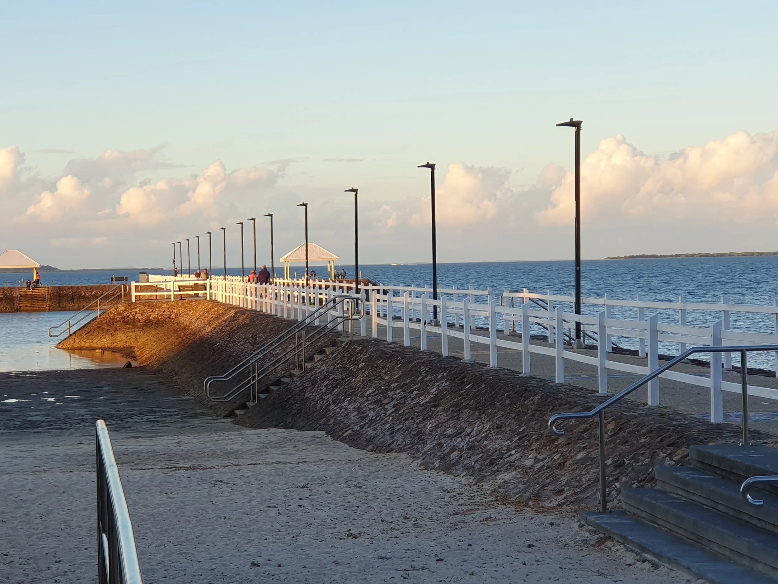 A seaside walkway with white fencing, black lampposts, steps leading down to a sandy area, and a small pavilion over the water, with a few people walking and relaxing on the pier.