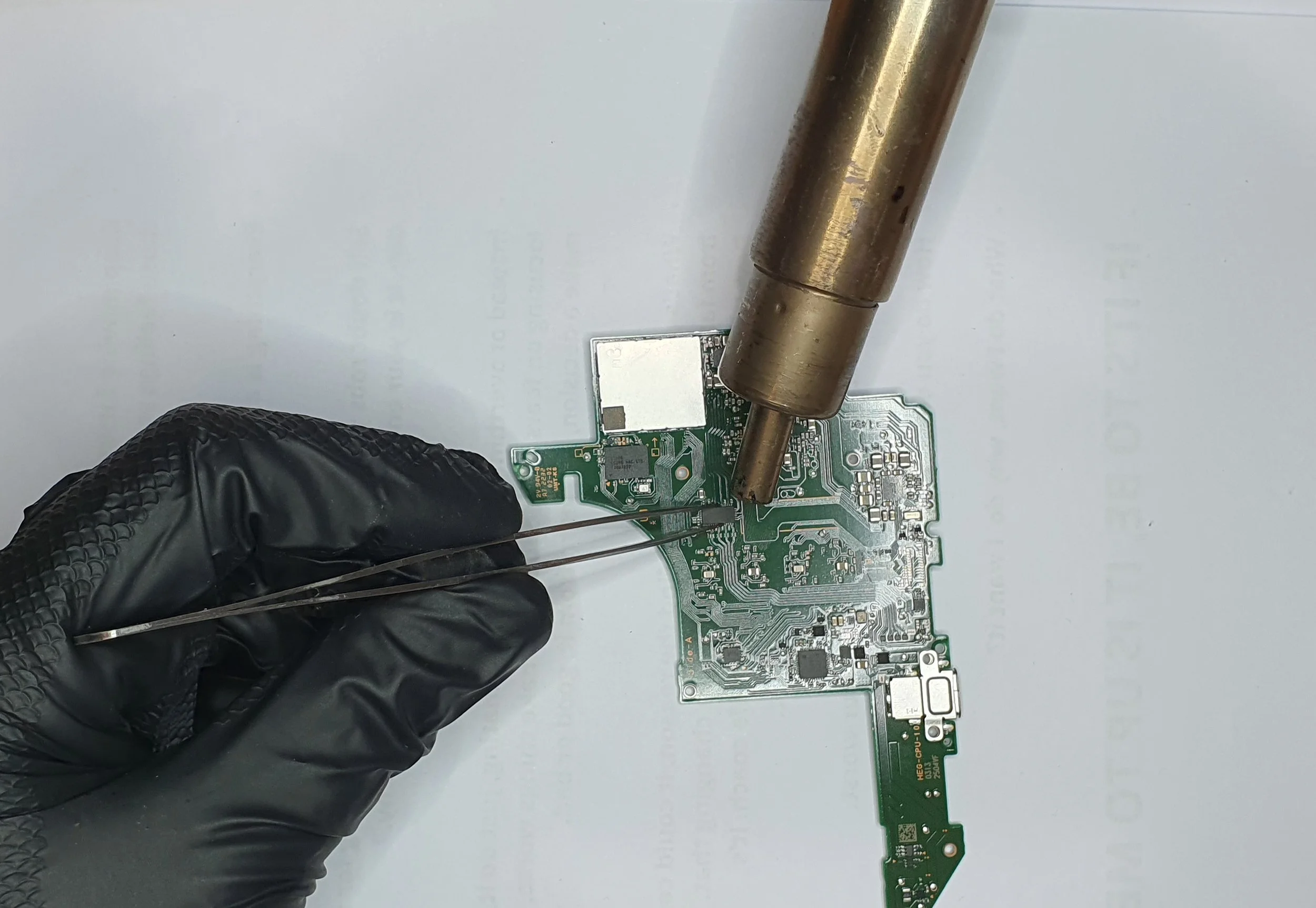 A person wearing black gloves uses a soldering iron to work on a green printed circuit board.