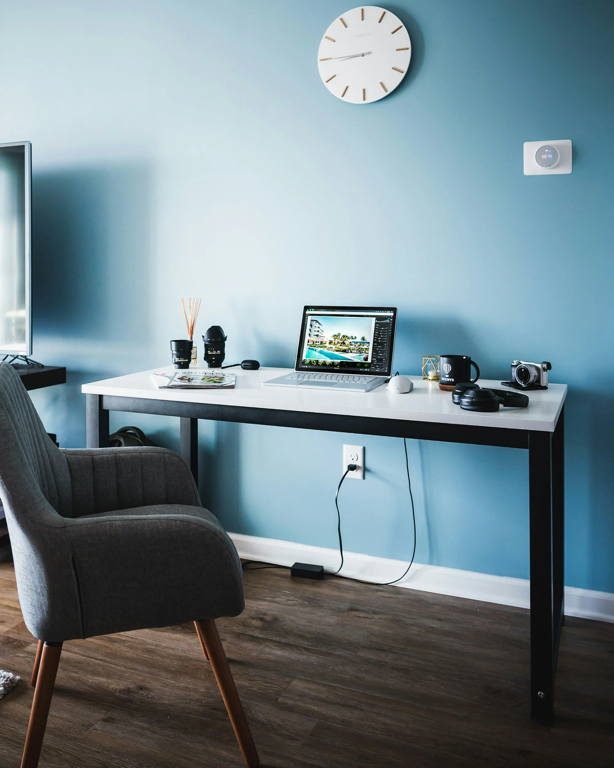 A workspace with a white desk, a gray upholstered chair, and various items including a laptop displaying a beach scene, a camera, headphones, a coffee mug, and office supplies. A blue wall with a large round clock and a digital thermostat is also visible.