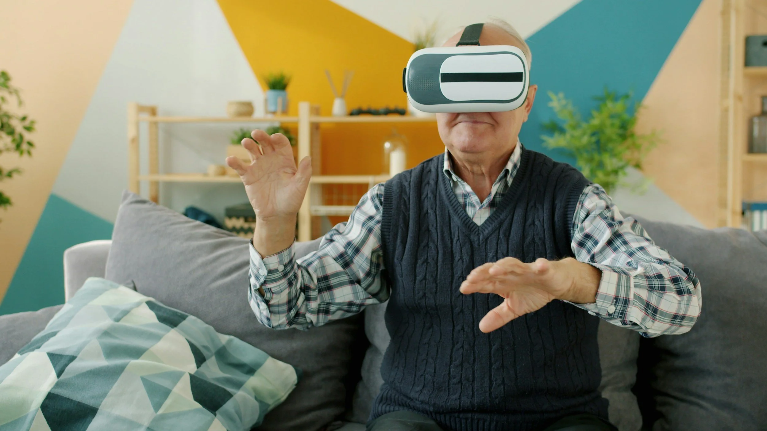 An elderly man sitting on a couch wearing a virtual reality headset and making hand gestures.