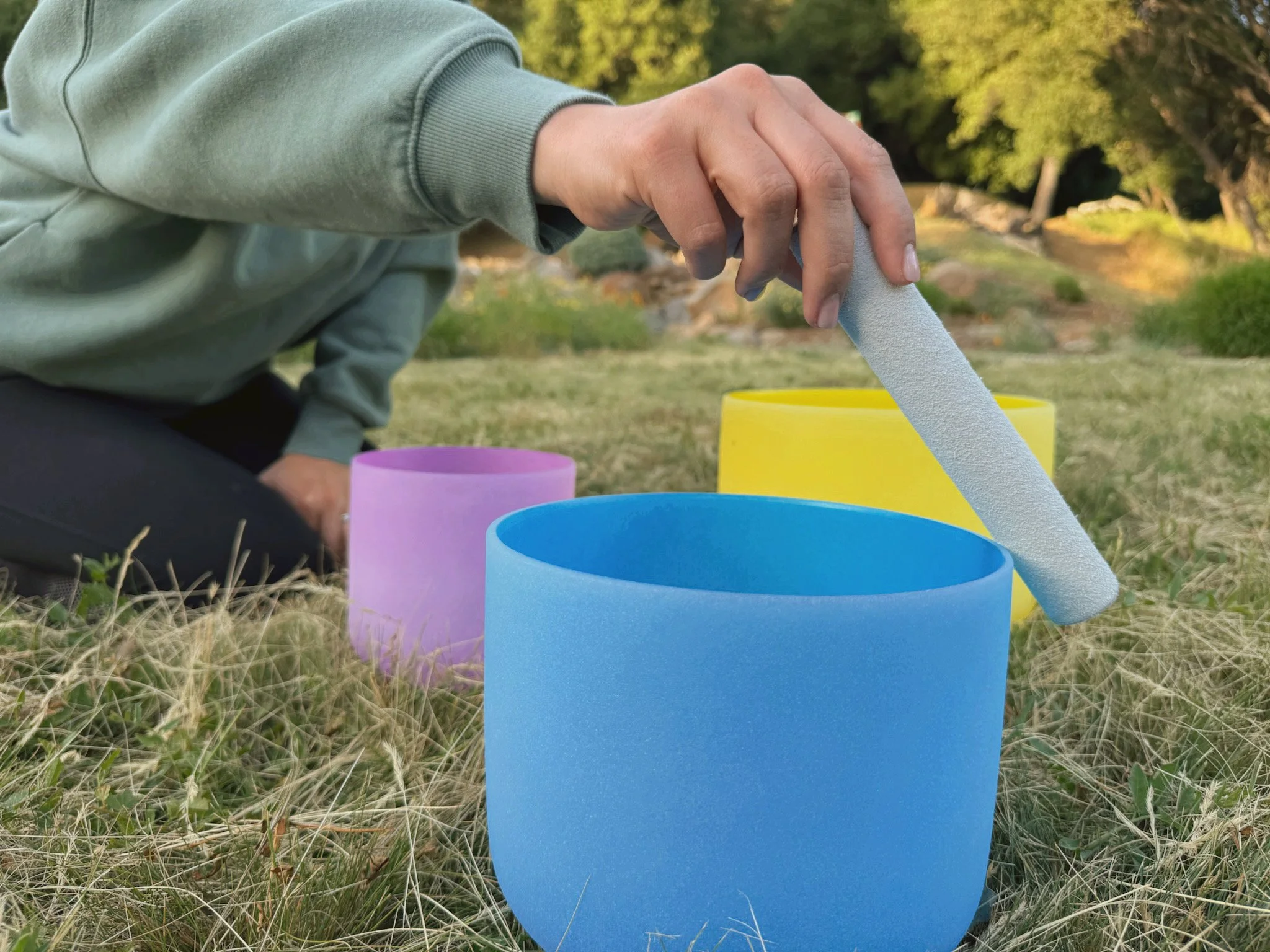 Close-up of a person's hand holding a gray foam noodle tube, with colorful plastic buckets nearby on grass, outdoors with trees in the background.