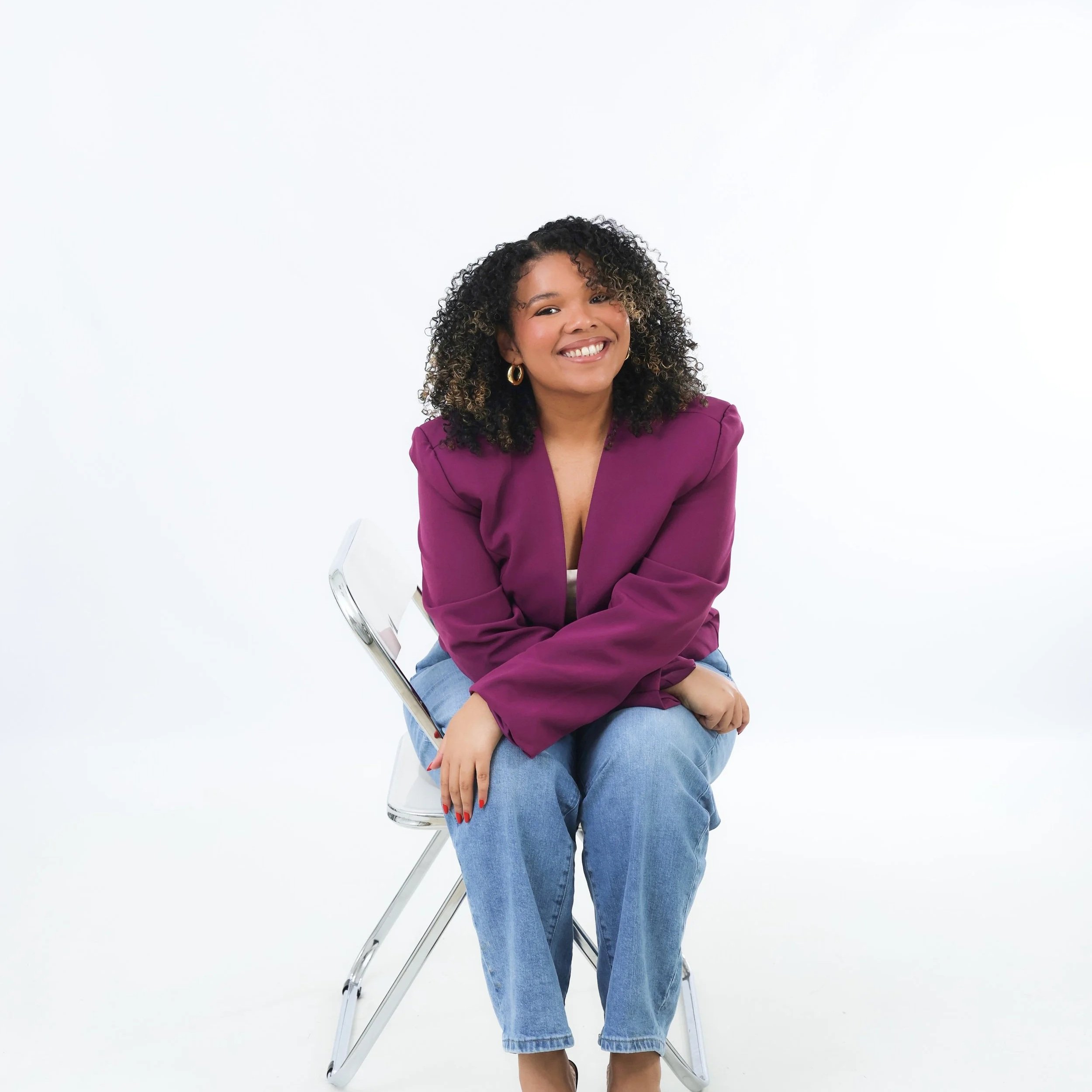 A woman with curly hair and gold hoop earrings smiling, sitting on a clear chair against a white background, wearing a purple blazer and blue jeans.