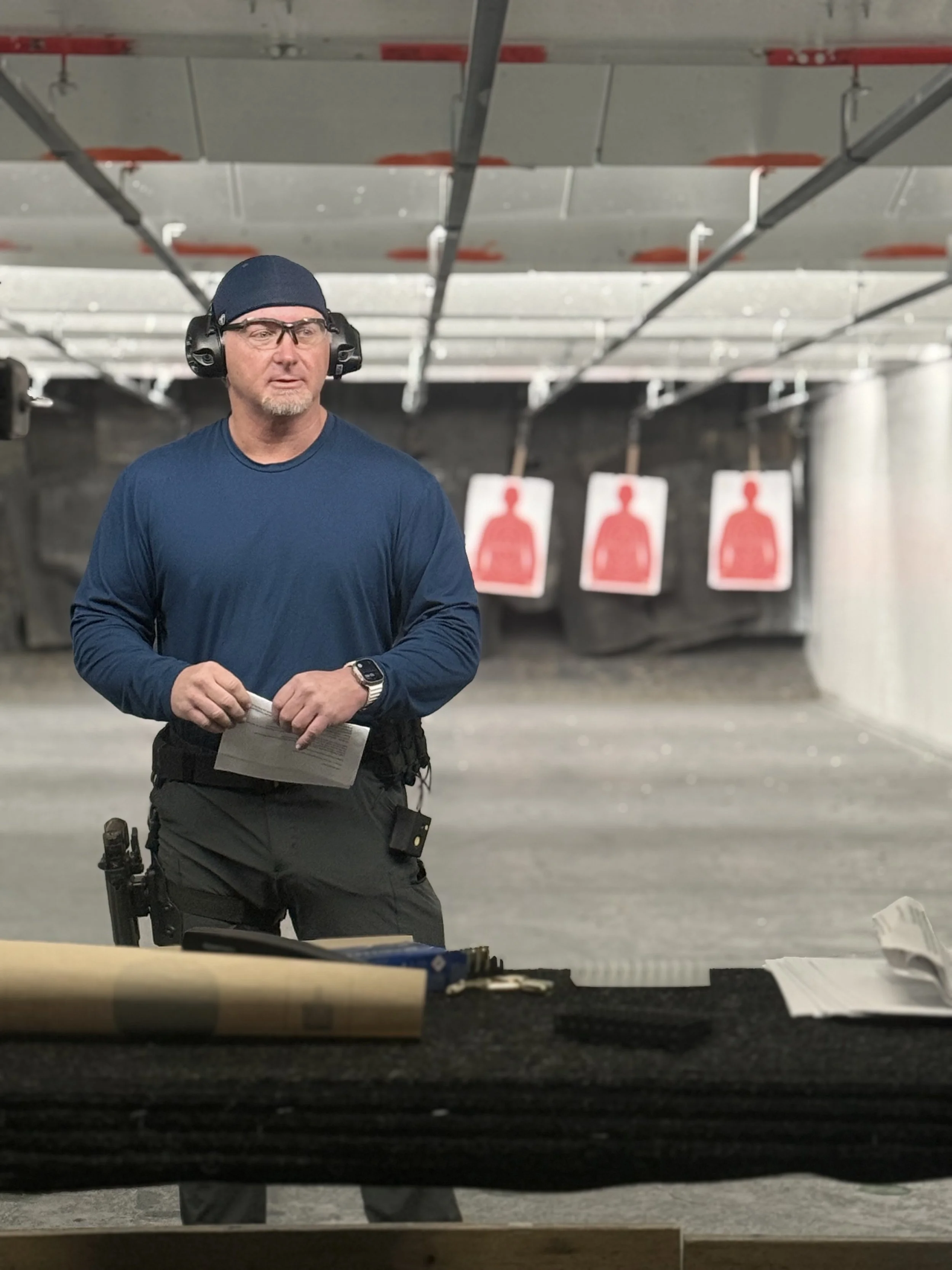 A man at an indoor shooting range wearing safety glasses, ear protection, a blue long sleeve shirt, and a cap, holding papers with three target silhouettes with red circles behind him.