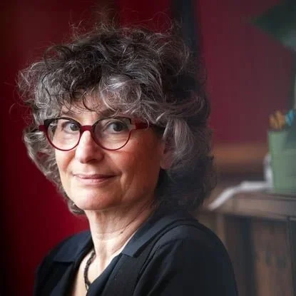 Woman with curly hair and red glasses smiling, wearing a black shirt, sitting indoors.