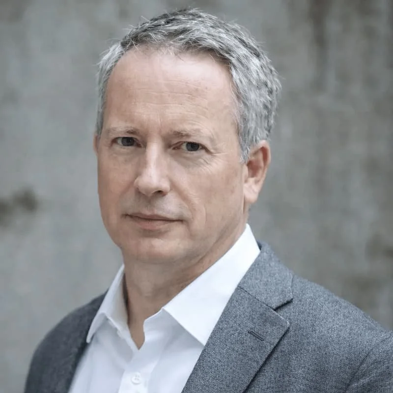 A man in a suit with a blue and white checkered shirt and a dark tie, smiling against a light gray background.