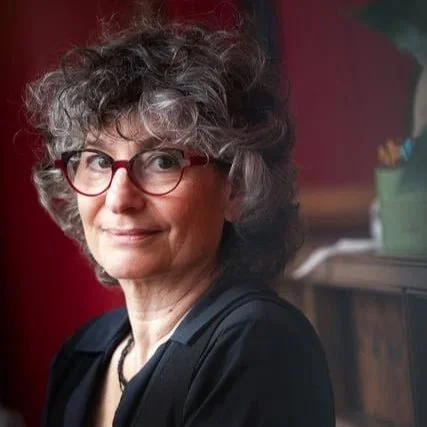 Woman with curly hair and red glasses smiling, wearing a black shirt, sitting indoors.