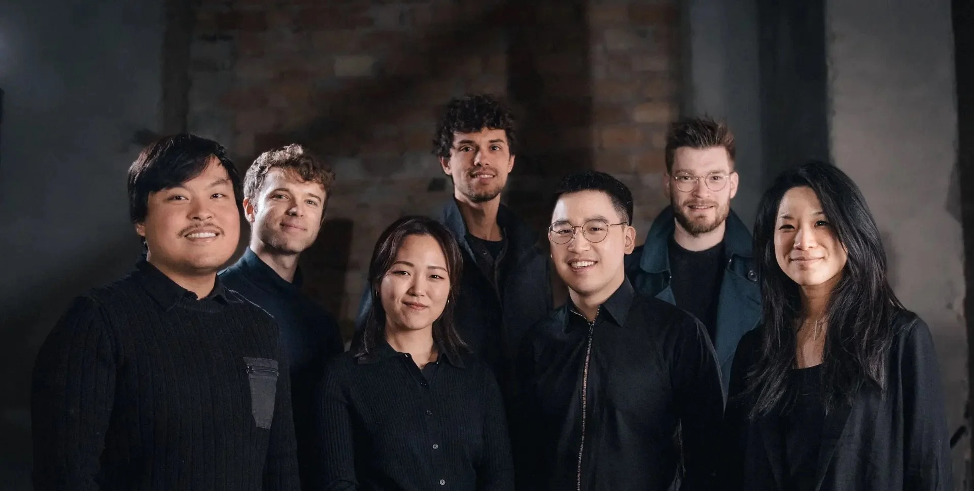 A group of seven people posing indoors, all dressed in dark clothing, standing against a blurred background.