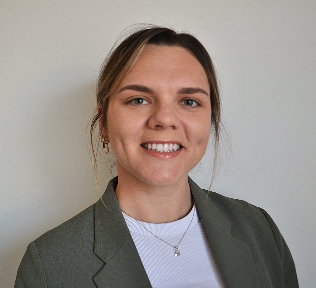 A woman with light brown hair, wearing a gray blazer and a white top, smiling at the camera against a plain light-colored wall.