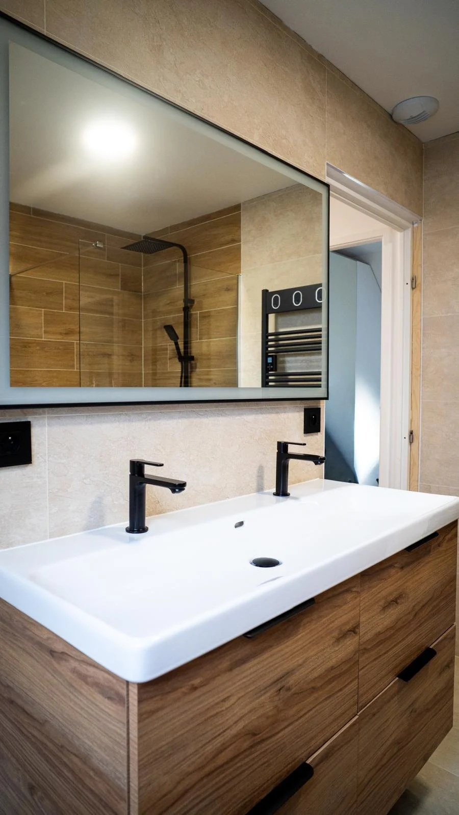Salle de bain avec meuble double lavabo en bois, miroir au-dessus, et douche avec parois en verre et douchette noire, murs en carrelage beige et bois.