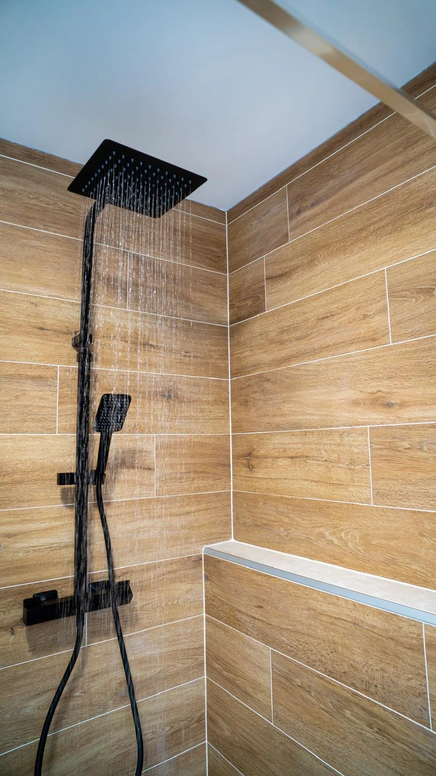 Douche à cabine avec une douchette noire au plafond et une autre à la main, murs en carreaux de bois, sur un mur de douche.